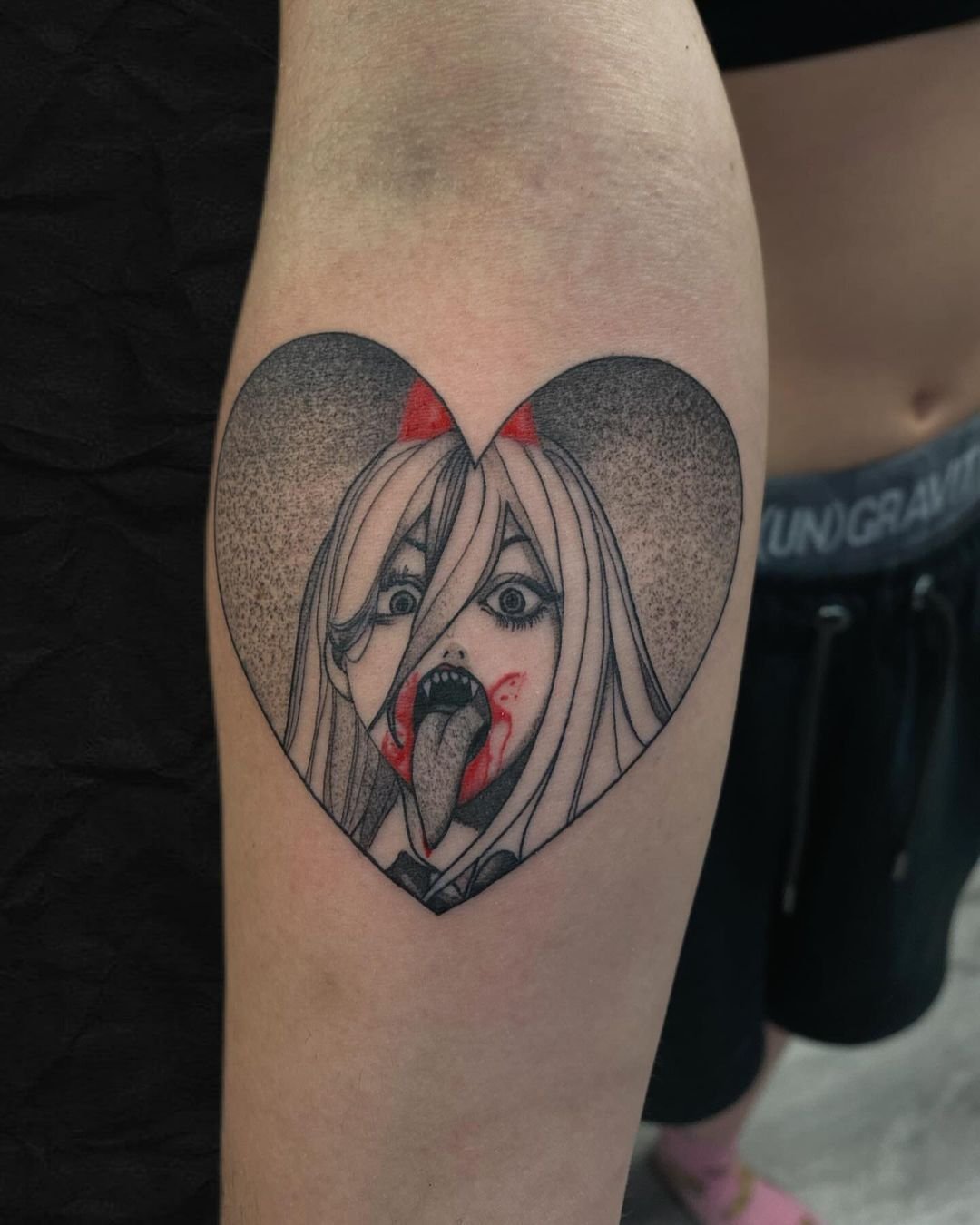 Tattoo of a cartoon character with long hair, sharp teeth, and open mouth inside a heart shape.