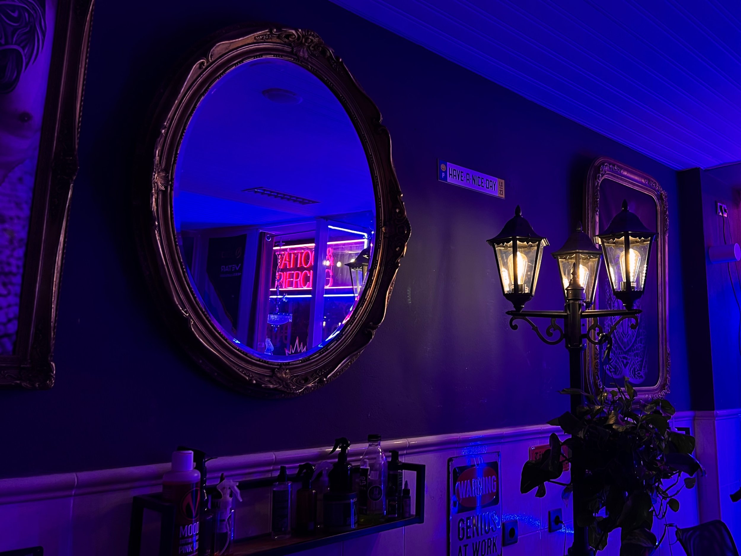 Dimly lit room with a round ornate mirror reflecting neon signs, a vintage-style street lamp, and wall signs including "Have a Nice Day" and "Genius at Work." The atmosphere features a mix of purple and blue hues, creating a moody ambiance.