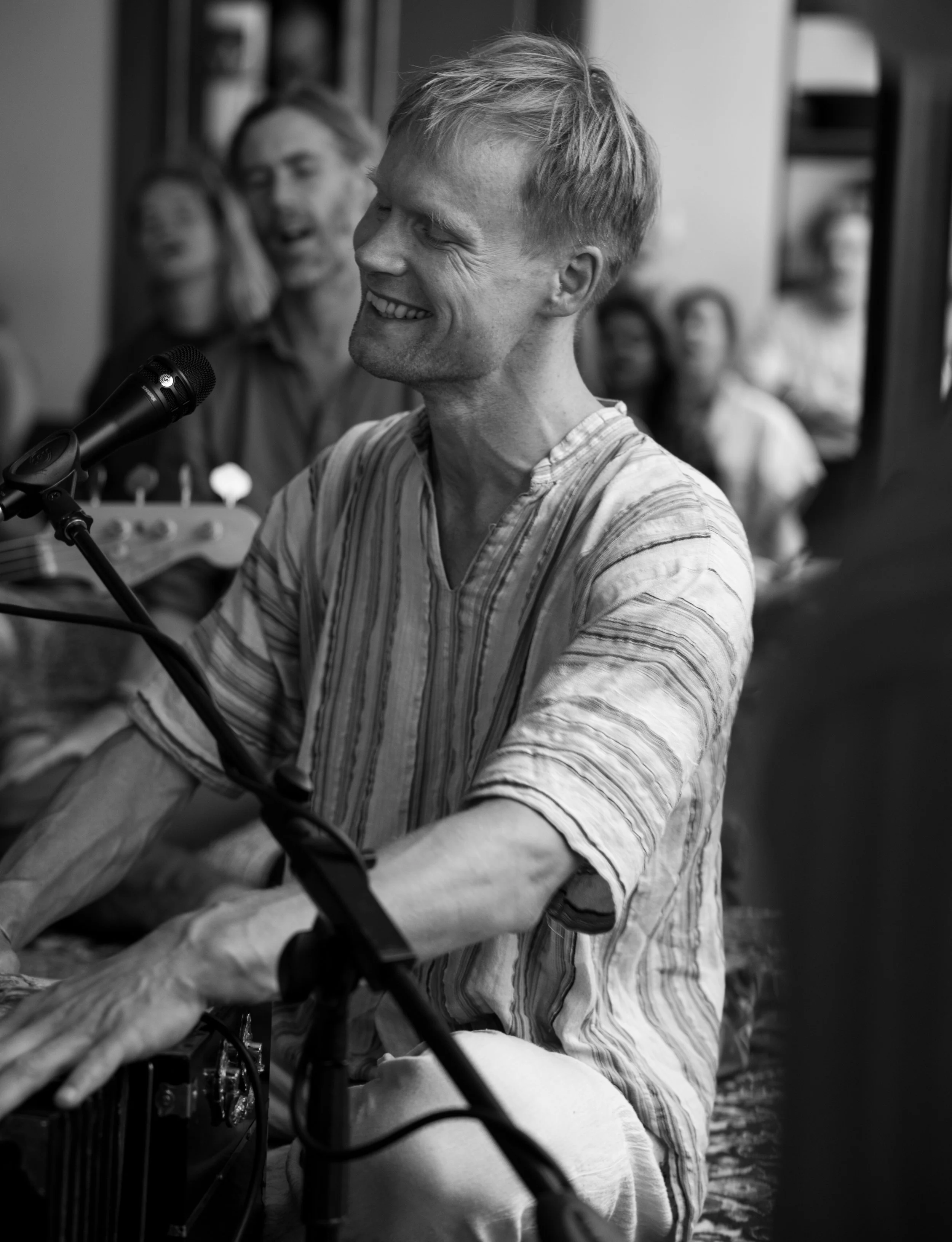 A young man with short, tousled hair smiling as he plays harmonium and sings into a microphone in a room with other singing people, some also smiling and laughing in the background.