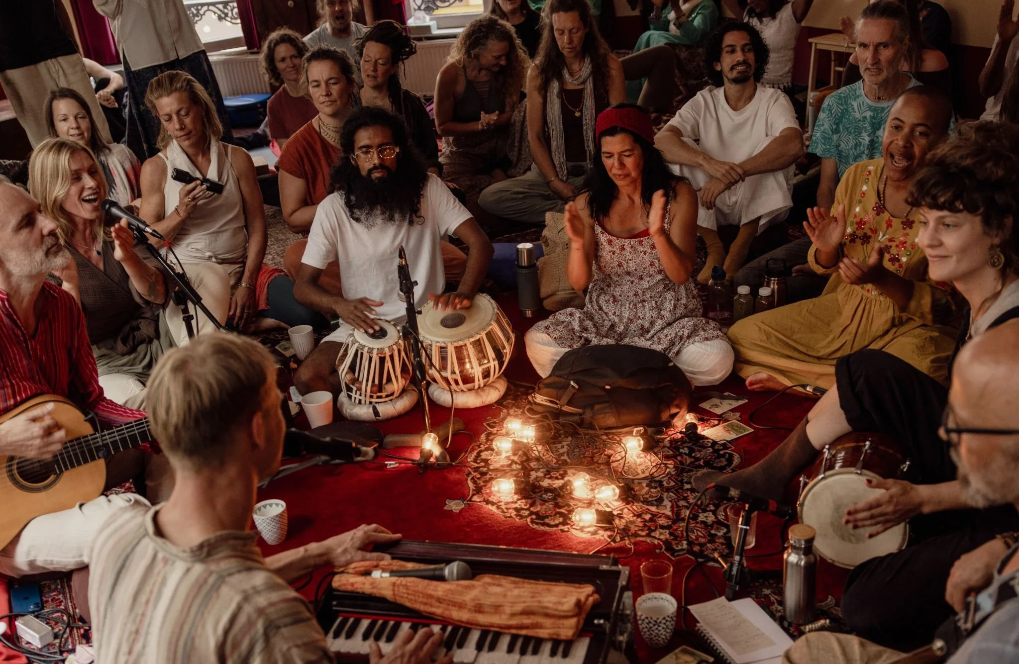 Large group of people gathered indoors for a lively musical kirtan jam session with microphones, guitars, hand drums, and harmonium.