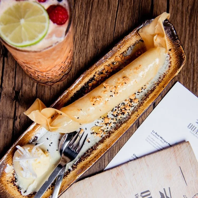 Ein cremiges Dessert mit Nüssen und Krokant, serviert auf einem langen, rechteckigen Teller, daneben ein Messer und Gabel. Im Hintergrund ein Glas mit einem Getränk, das Zitronenscheiben und Beeren enthält, auf einem Holztisch.