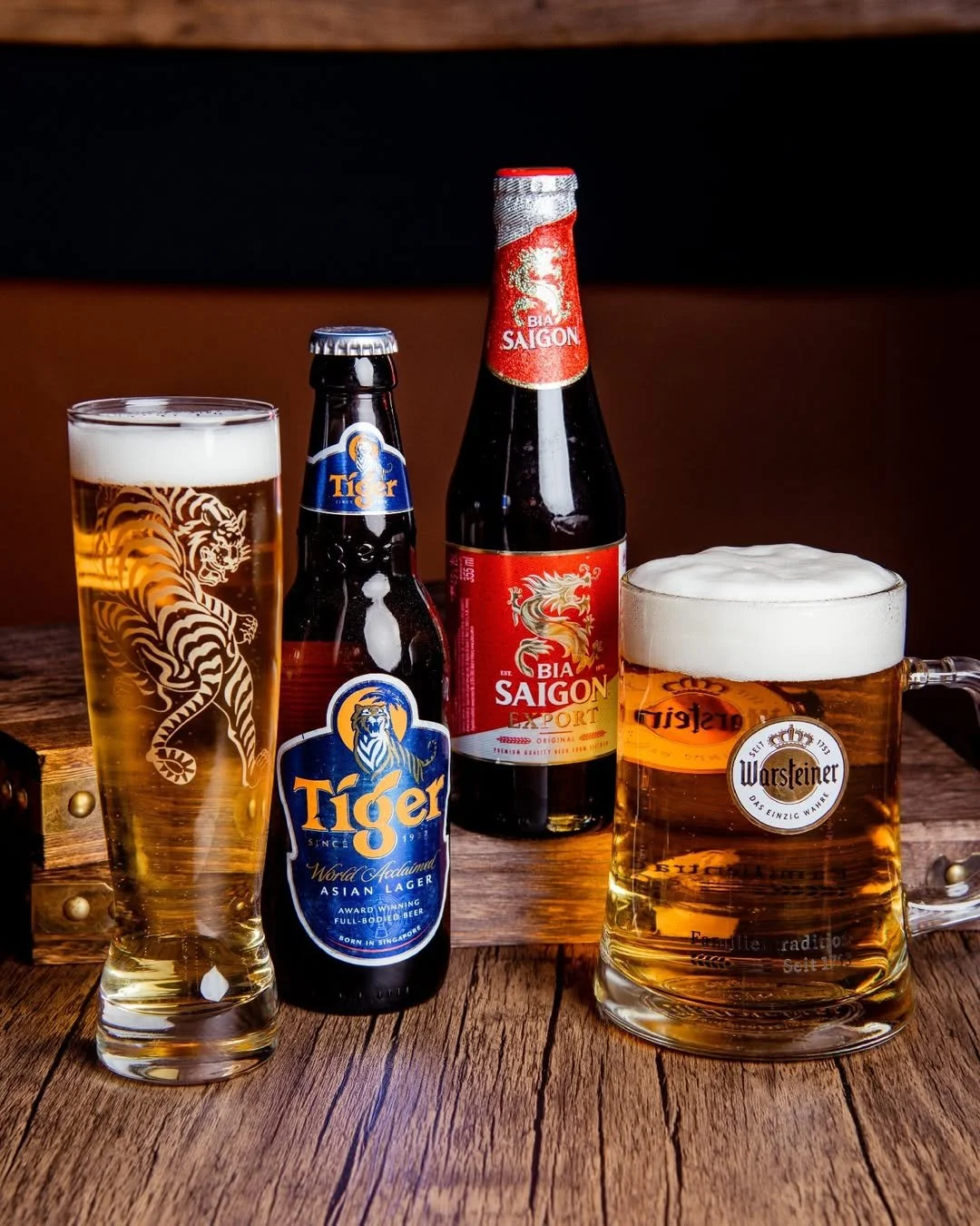 Vier Getränke, darunter ein Glas mit Goldbier, eine Flasche Tiger Beer, eine Flasche Bia Saigon Export und ein Krug Warsteiner Bier, stehen auf einem Holztisch.