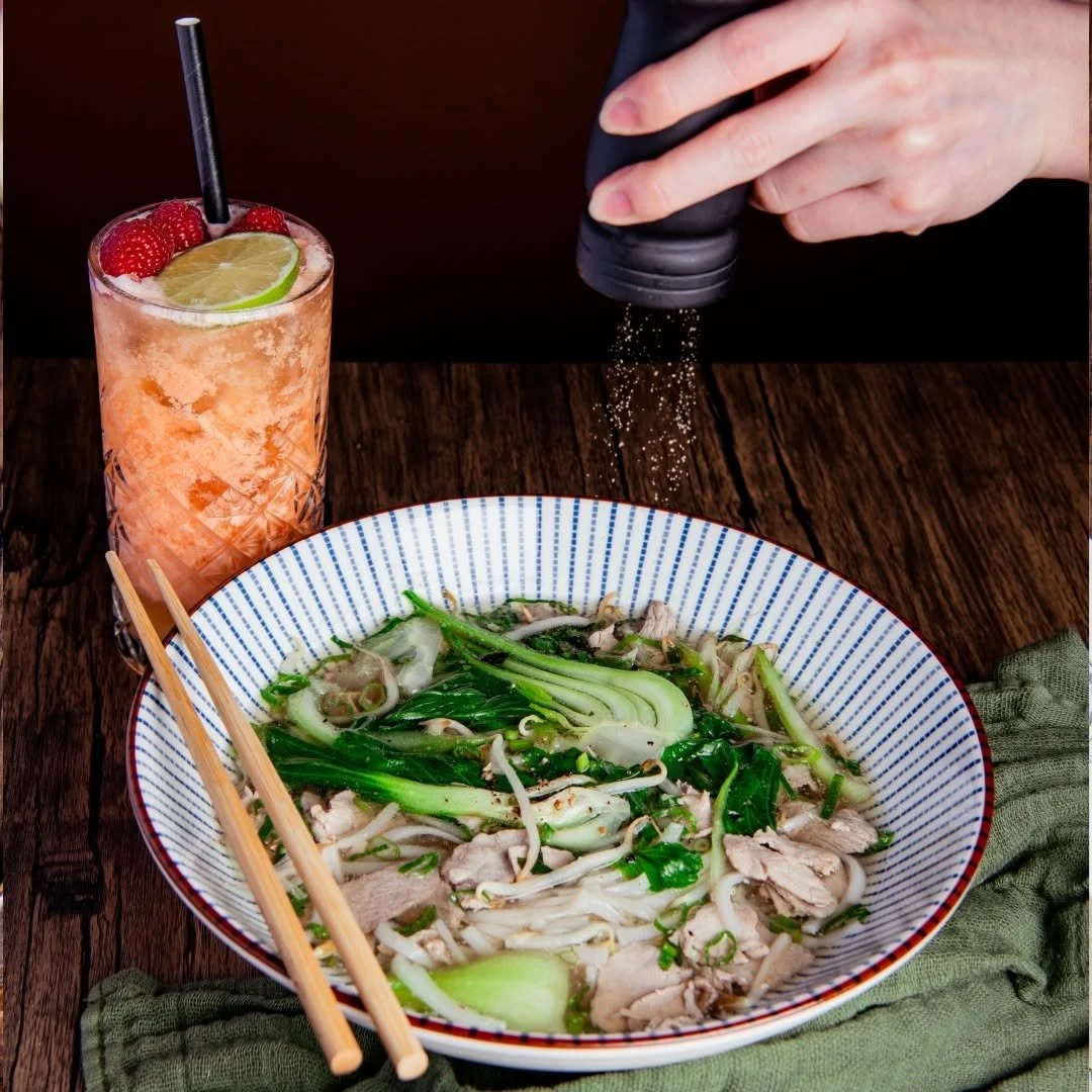Eine Schüssel mit Pho, vietnamesische Nudelsuppe, mit Hähnchenfleisch, Frühlingszwiebeln und Kräutern. Daneben eine Glas mit einer rosa Limonade, garniert mit Himbeeren und einer Limettenscheibe. Jemand streut Pfeffer über die Suppe.
