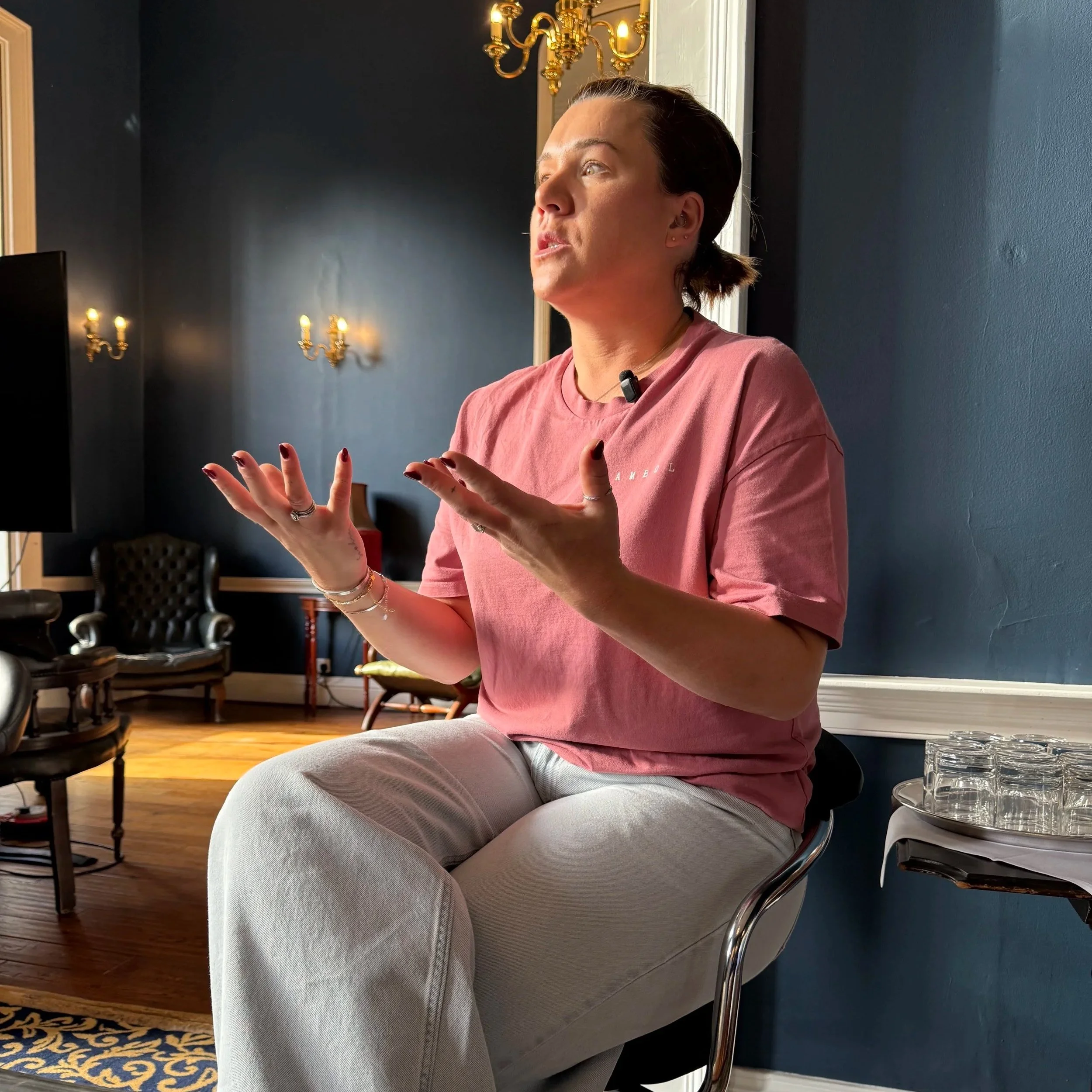 A person with short brown hair, wearing a pink shirt and multiple bracelets, sits on a silver chair with a black seat cushion. They are gesturing with their hands whie speaking. The background includes a dark blue wall, elegant gold wall sconces, and a mirror. There are also chairs and tables visible in the room.