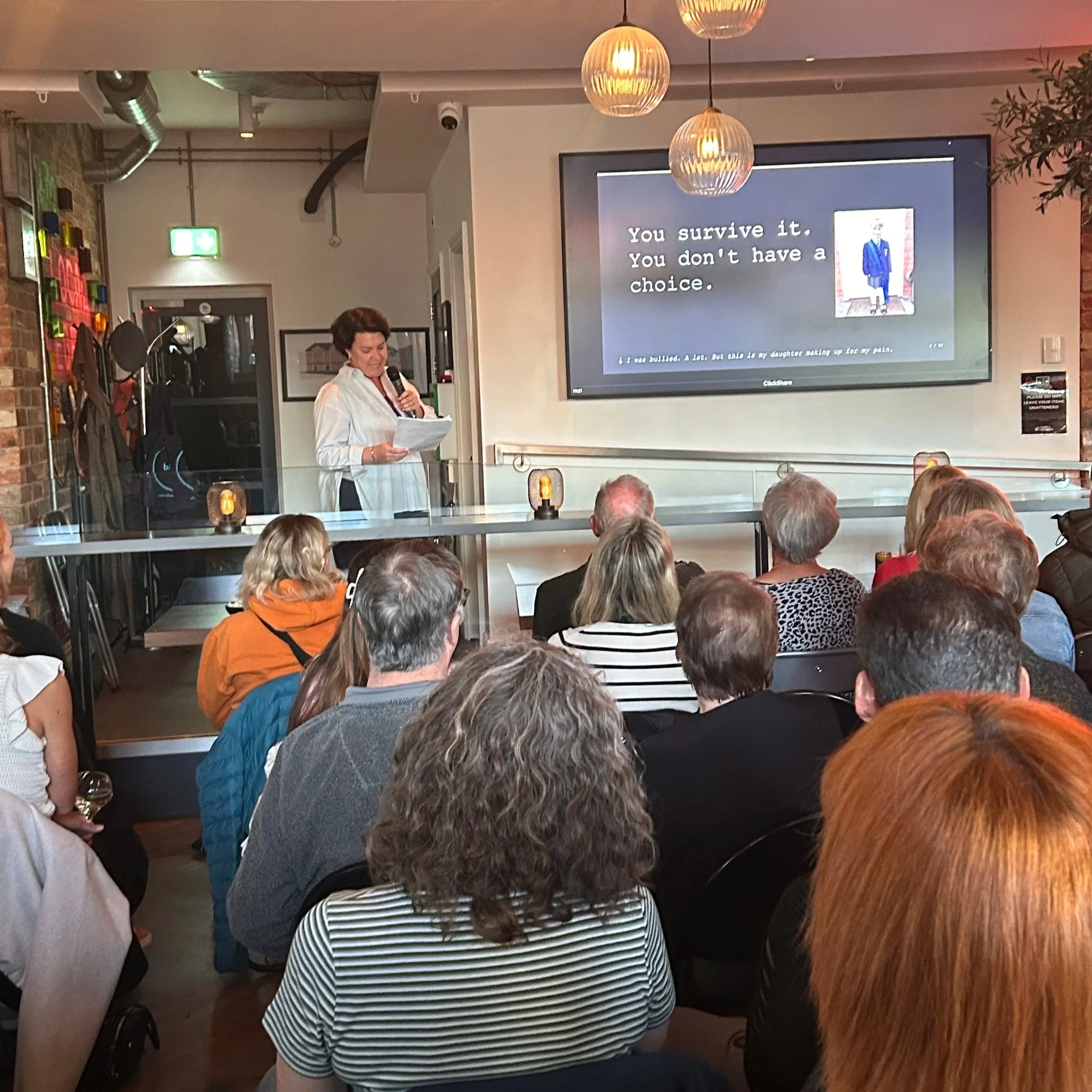 with dark hair wearing a white shirt is holding a microphone and reading from a paper, standing in front of an audience in a restaurant or cafe. A large screen behind her displays the text 'You survive it. You don't have a choice.' with a small cartoon image of a person in a blue suit and a brick wall background.