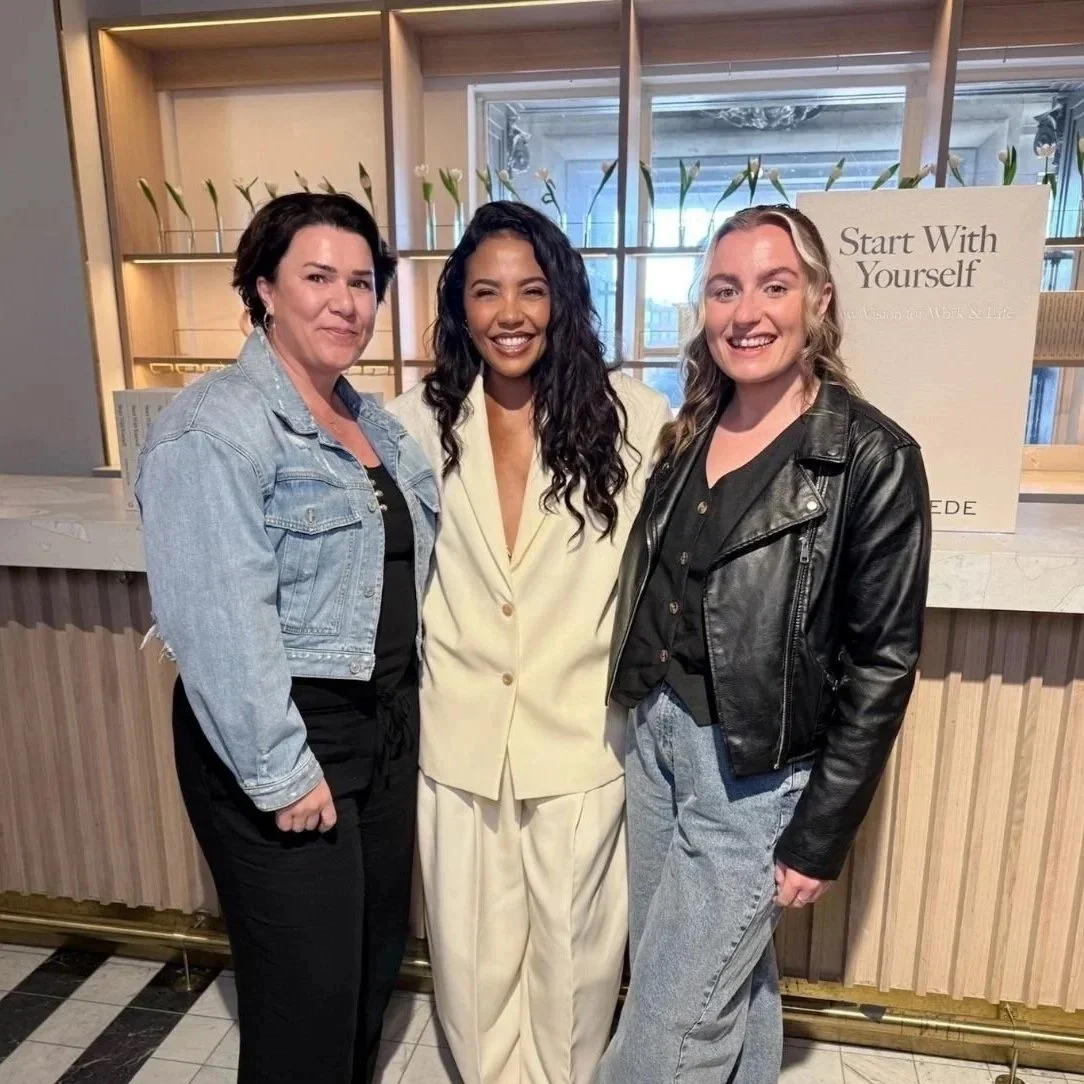 Three women standing together indoors in front of a wood and glass backlit display, smiling at camera.
