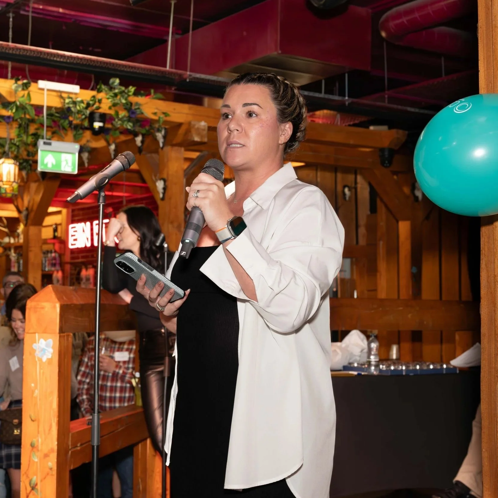 Jodie Horgan speaking into a microphone at an indoor event with wooden decor, a green exit sign, and a large blue balloon in the background.