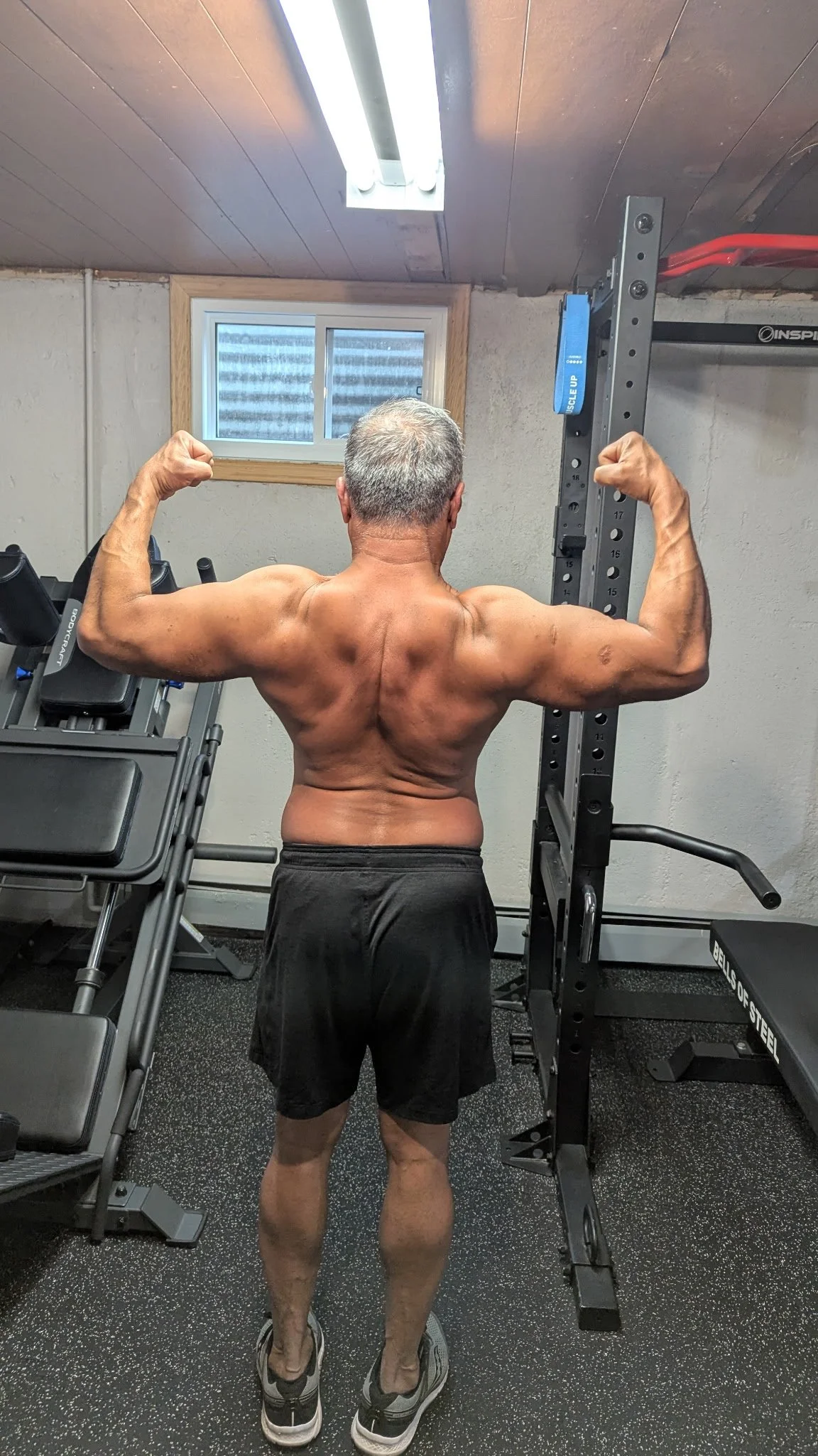 A muscular older man with gray hair, shirtless, flexing his biceps in a home gym. The photo is taken from behind, showing his back, arms, and legs. There are workout equipment and a small window in the background.