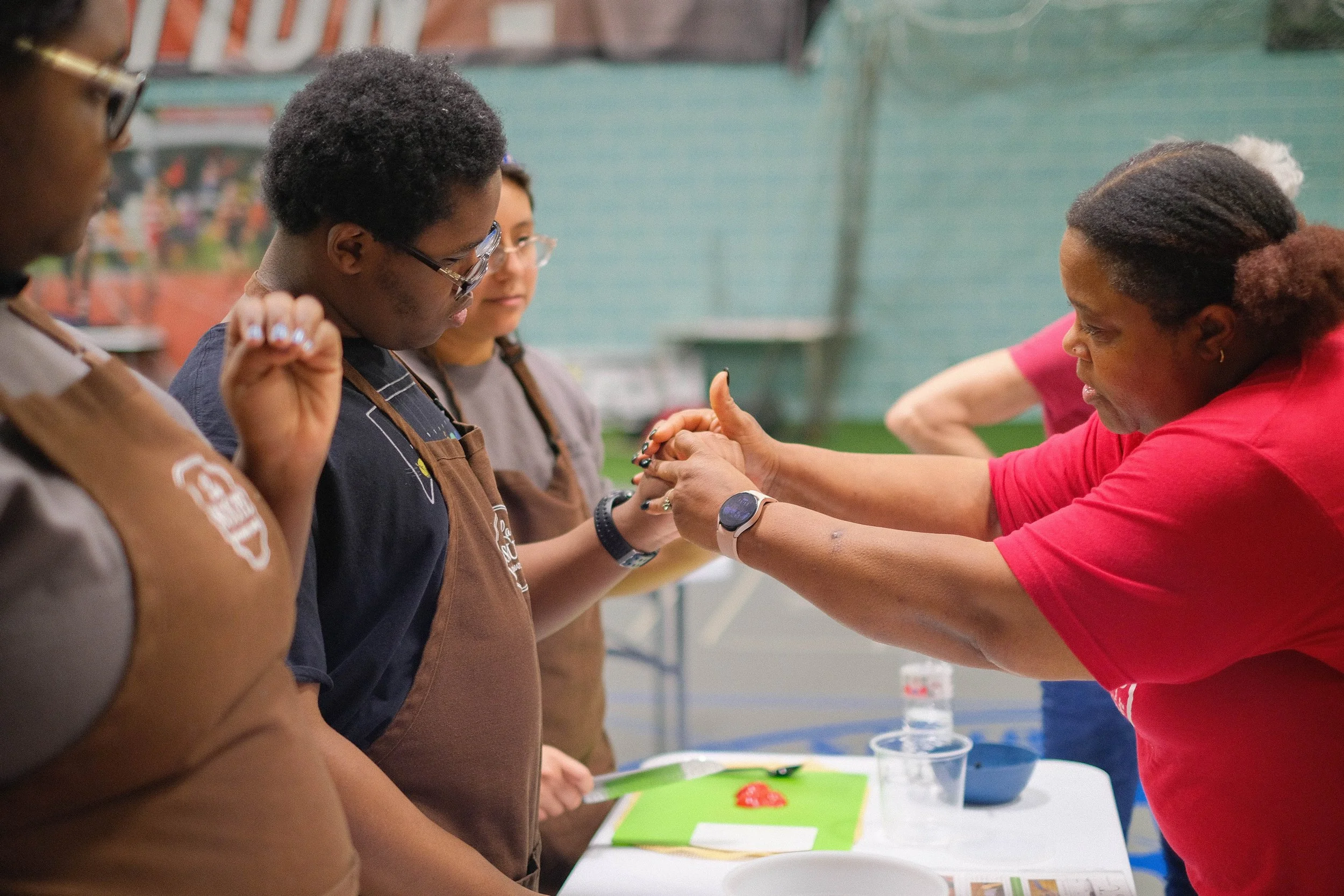 A woman in a red T-shirt shows a young boy in a brown apron how to properly position his fingers to hold a knife.