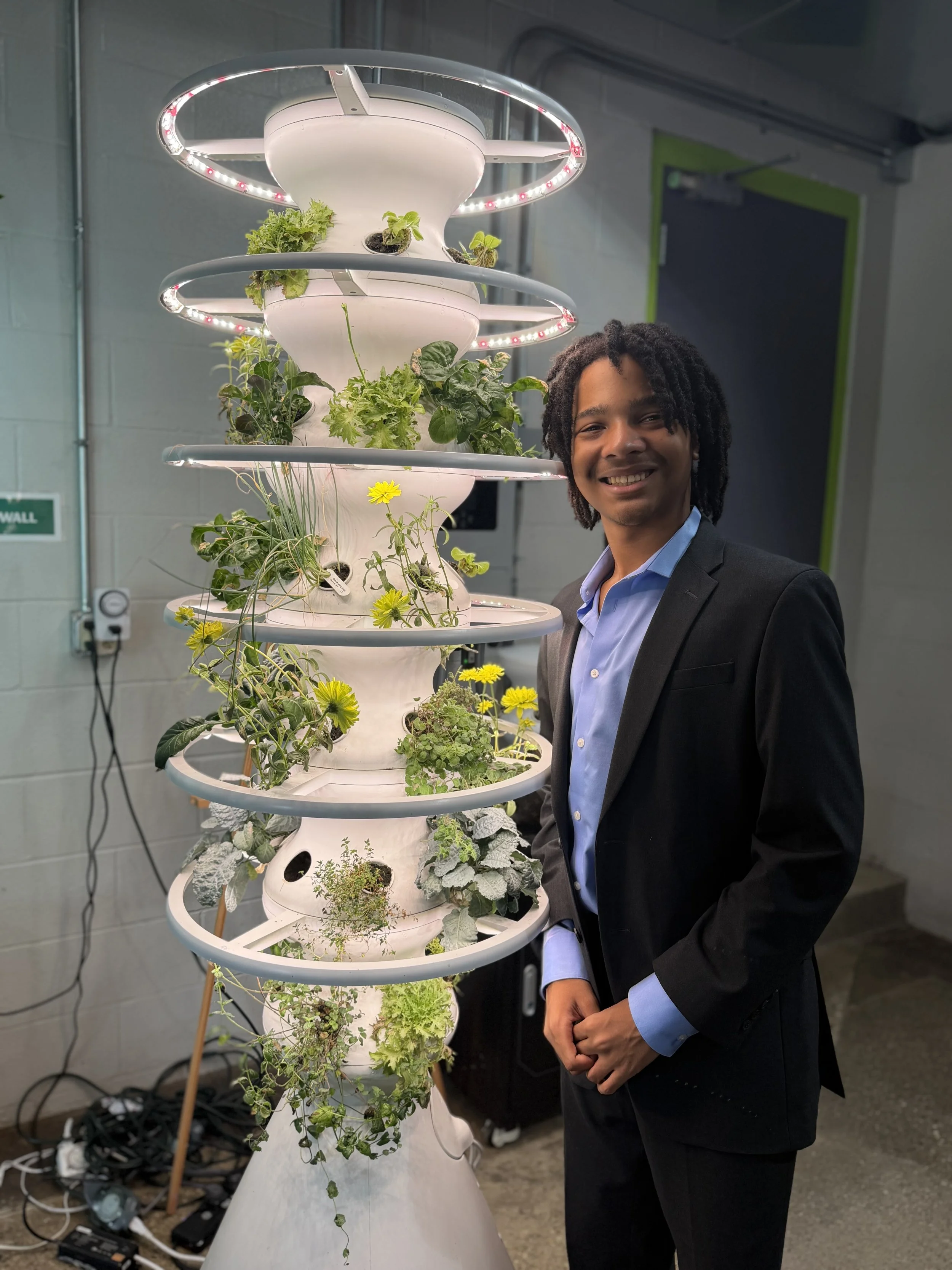 A young man in a blue dress shirt and a black suit stands by a white, futuristic tower planter that has green, leafy plants and round yellow flowers sprouting from it.
