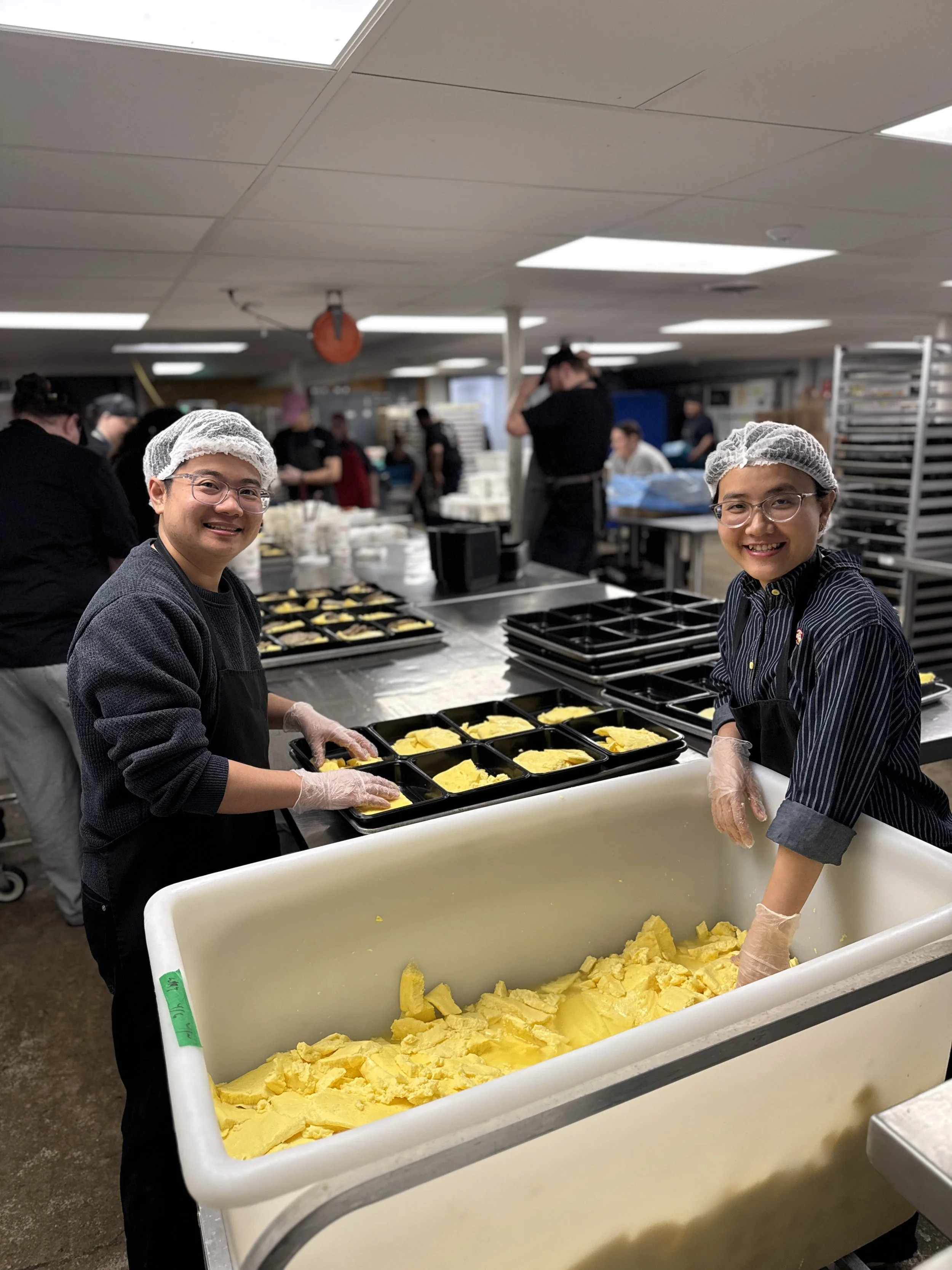 A young man and a young woman wearing hair nets and aprons smile as they put food into small black pans.