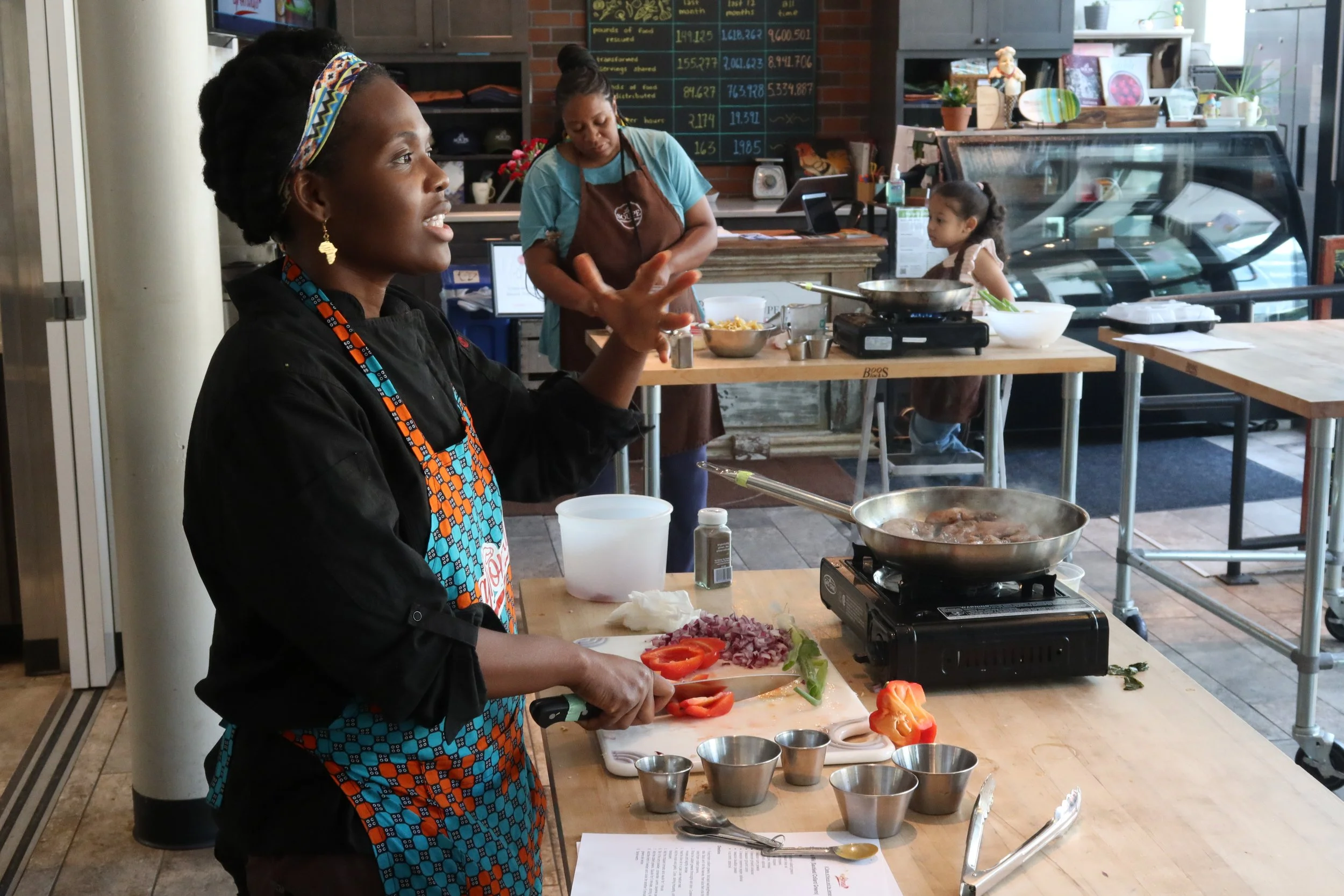 A woman in an apron stands at a wooden table, holds a kitchen knife, and prepares to cut vegetables.
