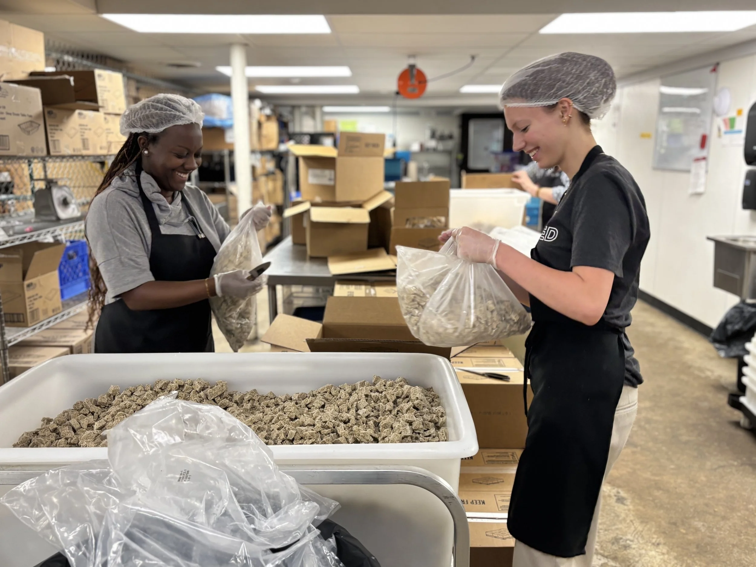 Two young women in hair nets and aprons put ingredients into clear plastic bags.