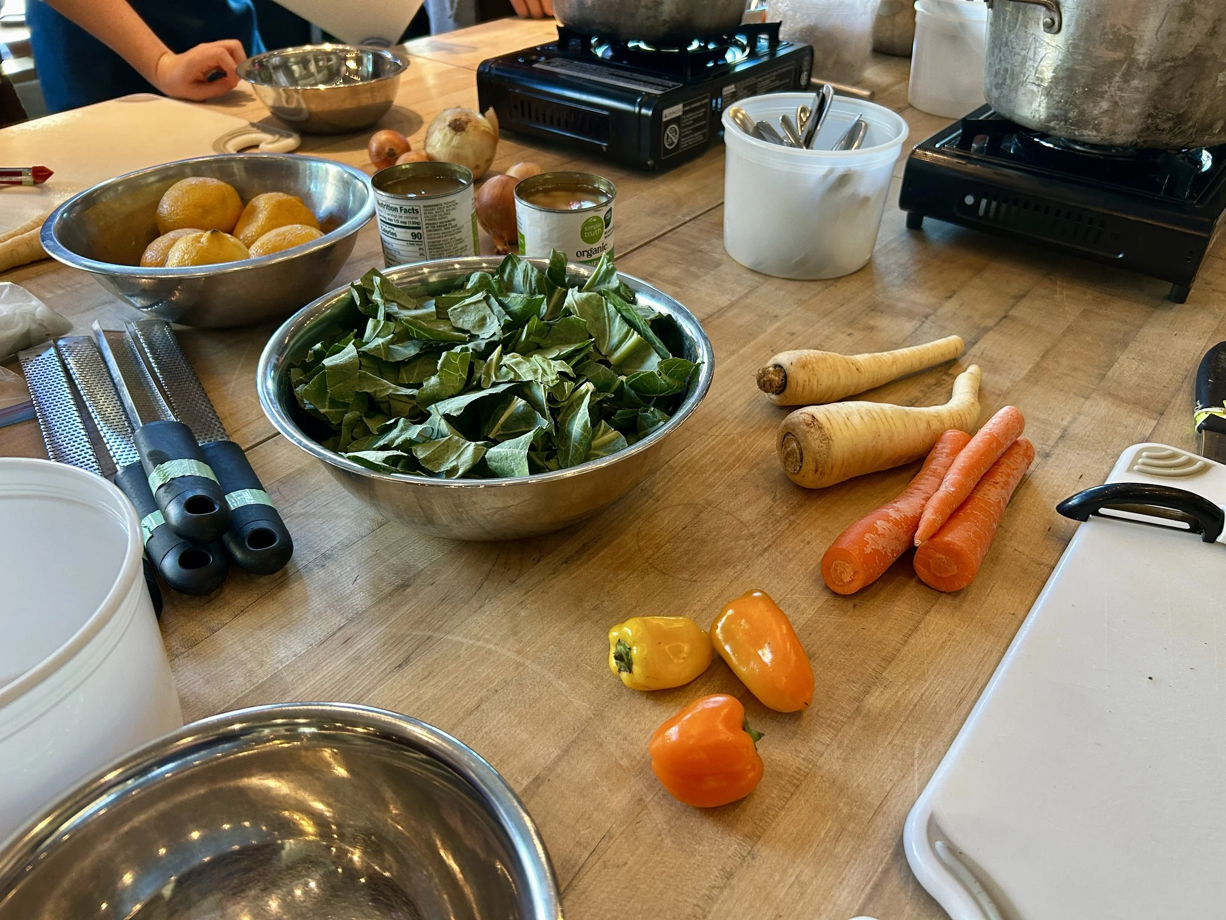 Bright yellow lemons and leafy green vegetables sit on a wooden table in silver bowls. They're surrounded by other red, orange, and yellow veggies like peppers and parsnips.