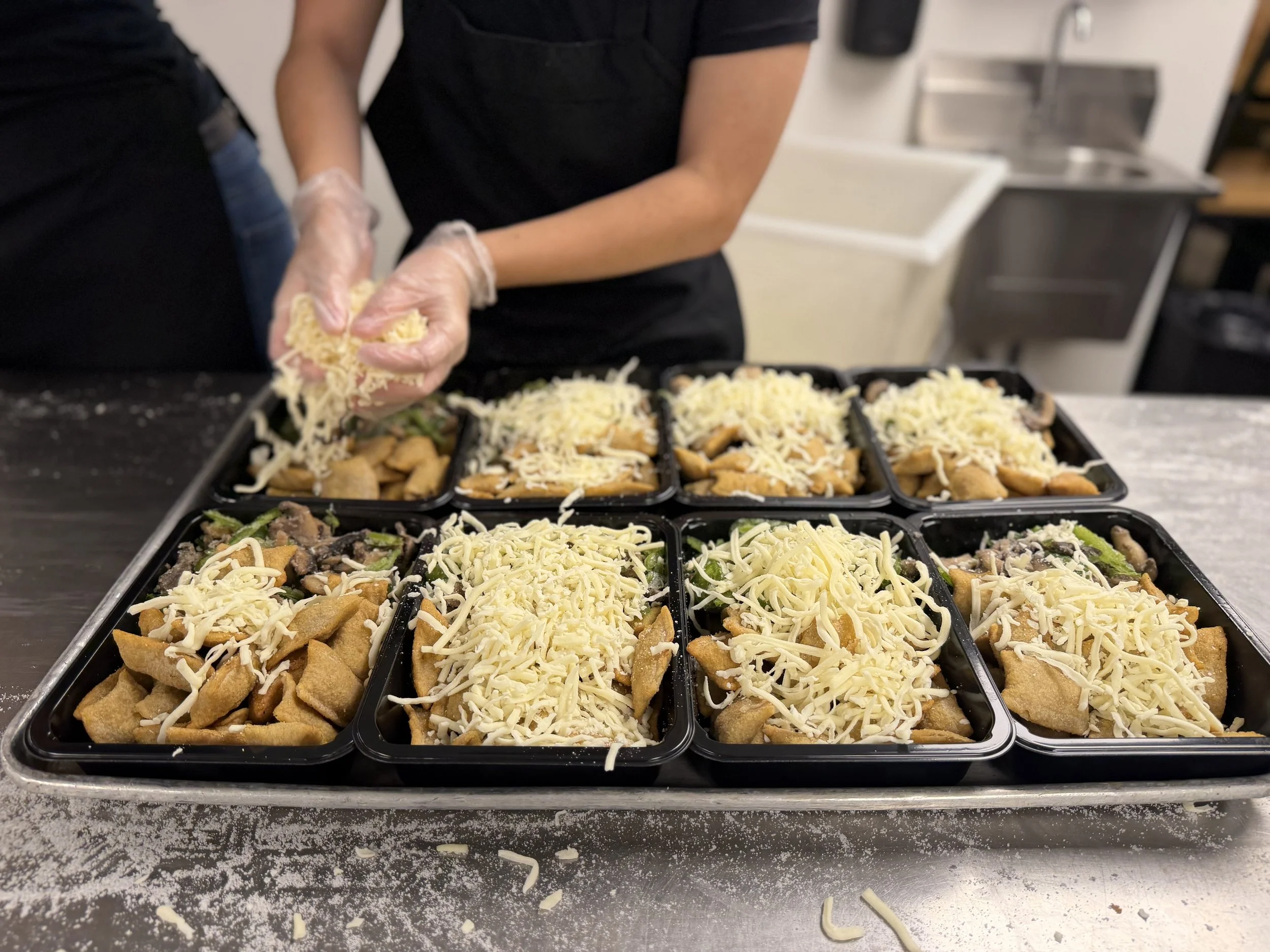 A person wearing kitchen gloves sprinkles shredded, light yellow cheese on tiny pans filled with pizza rolls and green veggies.