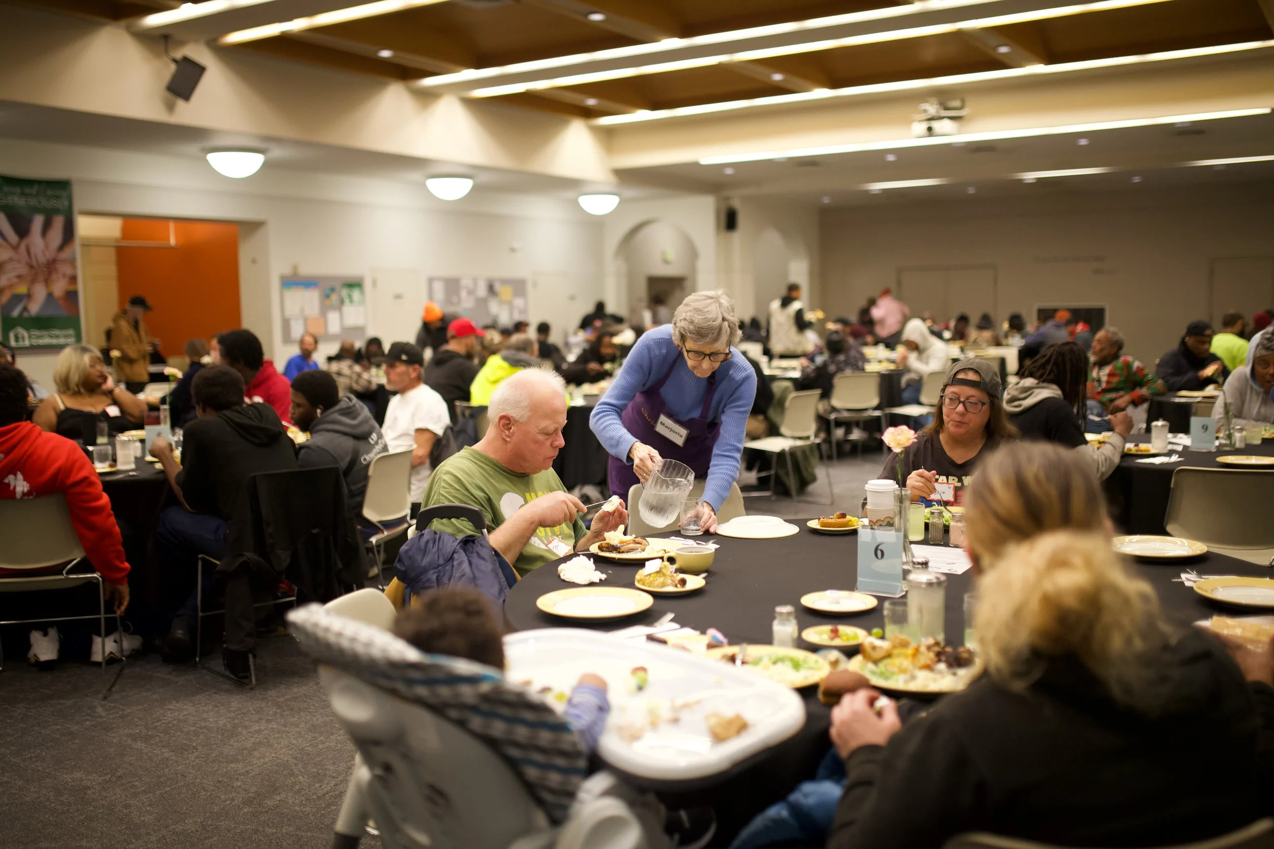 A small crowd of people gathers in a bsement for dinner. Tables are round with black table cloths, and an older woman in a bright blue sweater refills an older man's water glass with care.