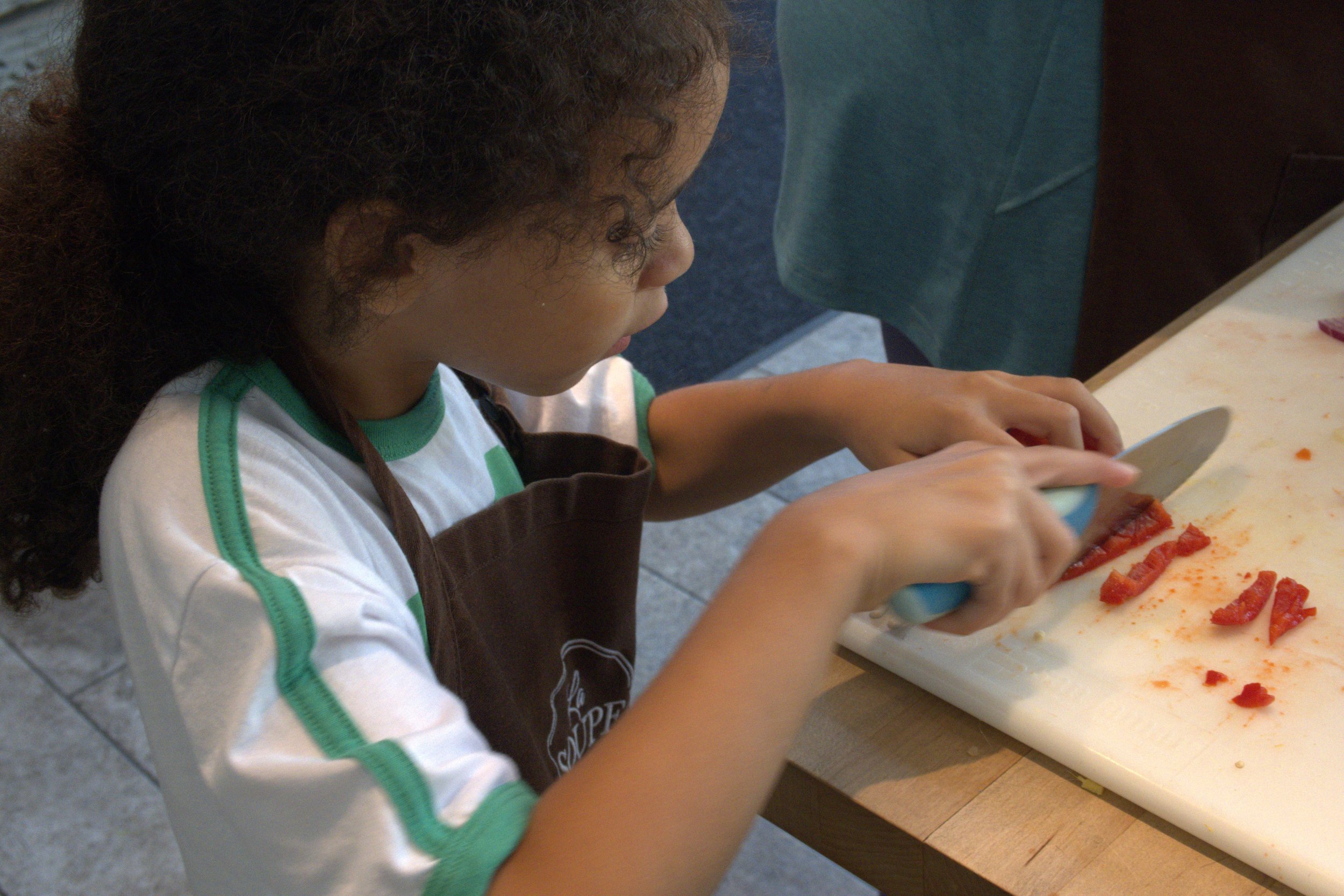 A young child using a knife to cut a red bell pepper on a cutting board.