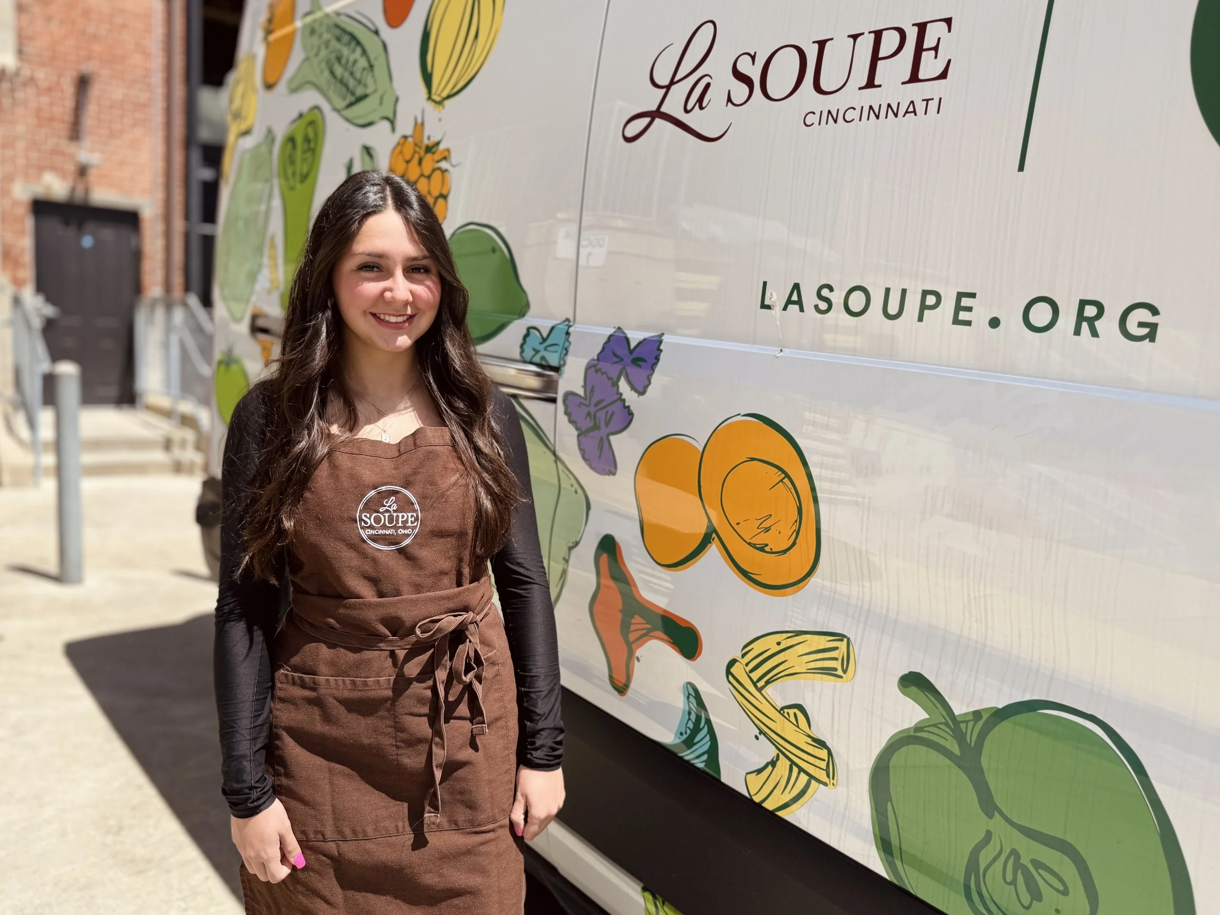 La Soupe Volunteer, Laney Sgantas, stands next to our Whole Foods delivery van in a brown La Soupe apron at our rescue kitchen in Walnut Hills, Cincinnati.