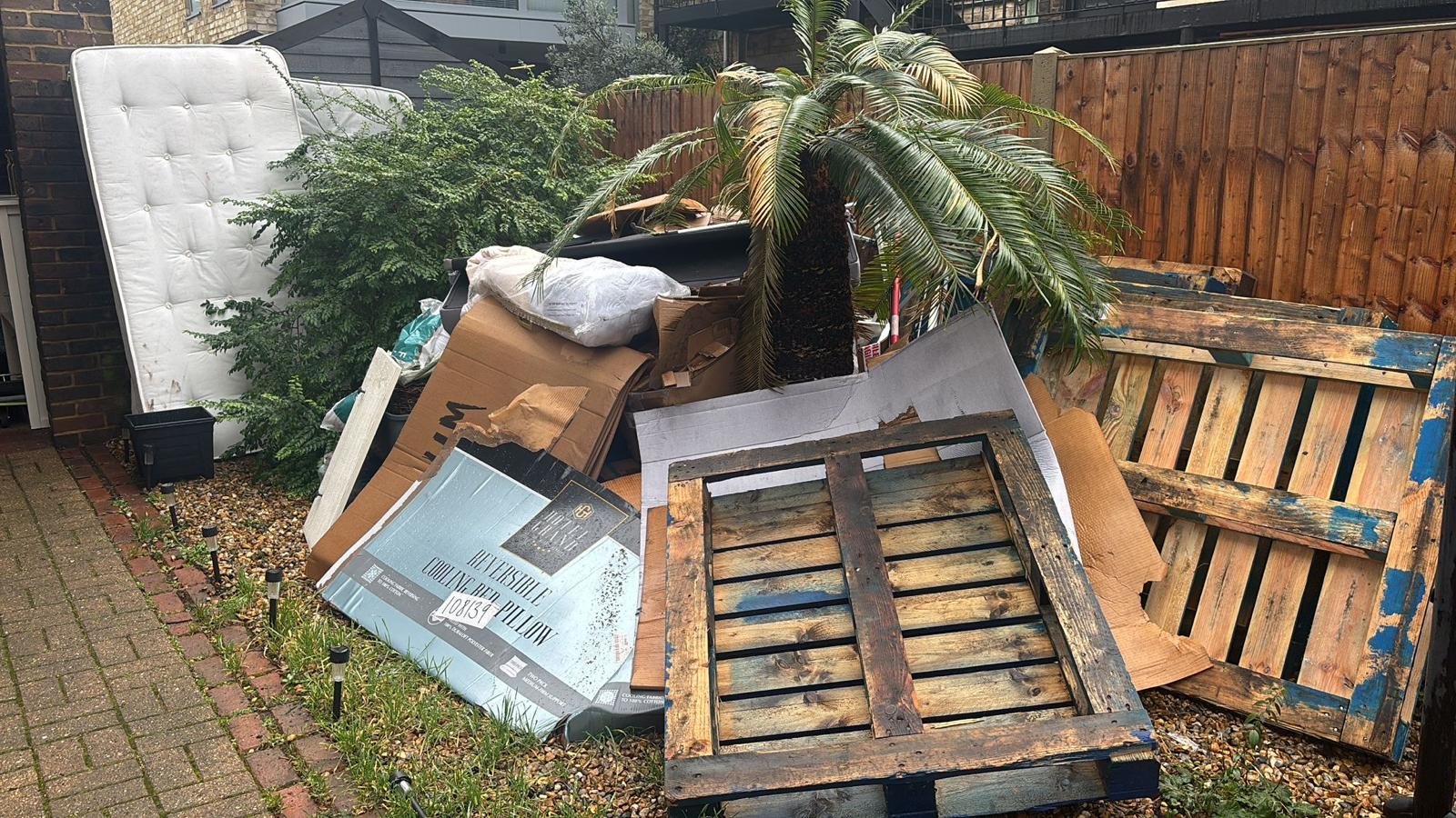 Garden Clearance in Clapham, clearing discarded furniture and yard debris, including a wooden pallet, a large palm tree, cardboard boxes, a mattress, and other miscellaneous items, piled against a wooden fence in a backyard.
