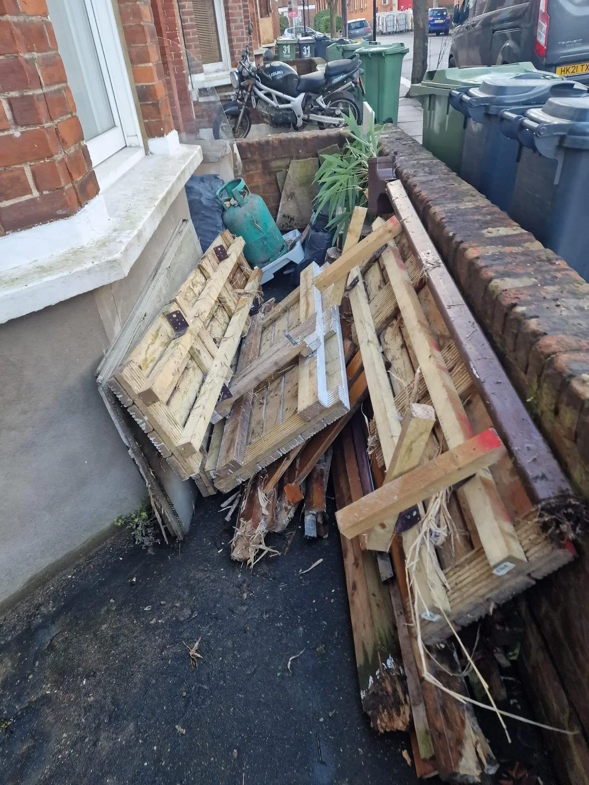 Garden Clearance in Clapham clearing a disorganized wooden pallets, a plant, a gas container, and some trash next to a brick wall and parked motorcycles on a city sidewalk.