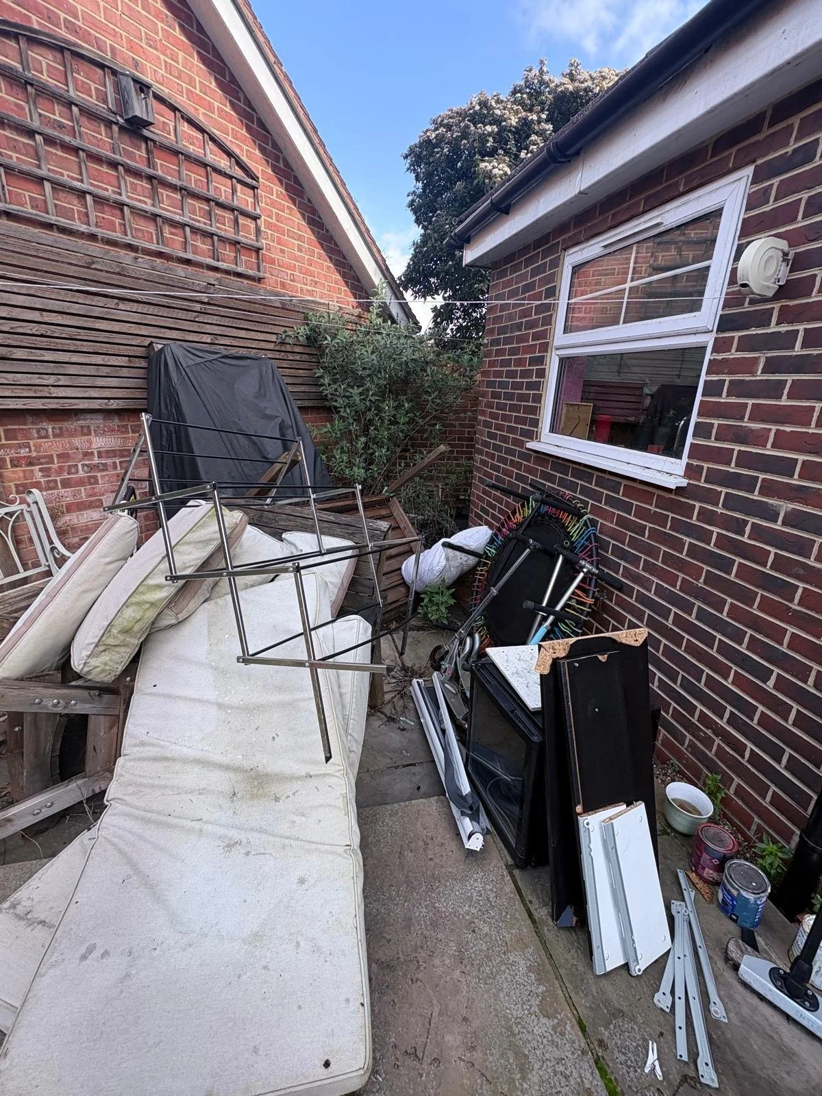 Garden Clearance in Clapham clearing a cluttered backyard with a mattress, metal furniture frame, scattered metal parts, a small trampoline, a small tree, and various household items next to a brick house.