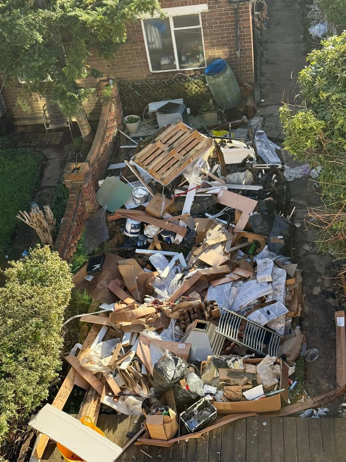 A backyard filled with construction debris and trash, including cardboard, wood, plastic, and metal materials, scattered across the ground surrounded by trees and a brick wall.