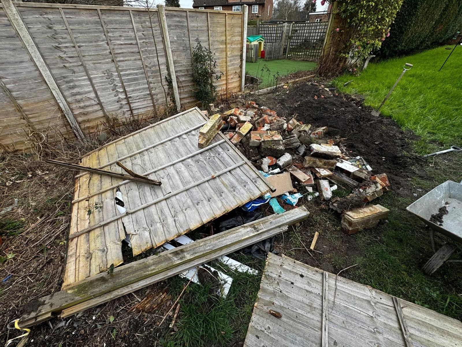 Garden Clearance in Clapham,  a backyard garden area with a collapsed wooden shed, scattered bricks, a pile of debris, a wheelbarrow filled with dirt, and gardening tools, enclosed by wooden fences and next to a grassy area with trees.