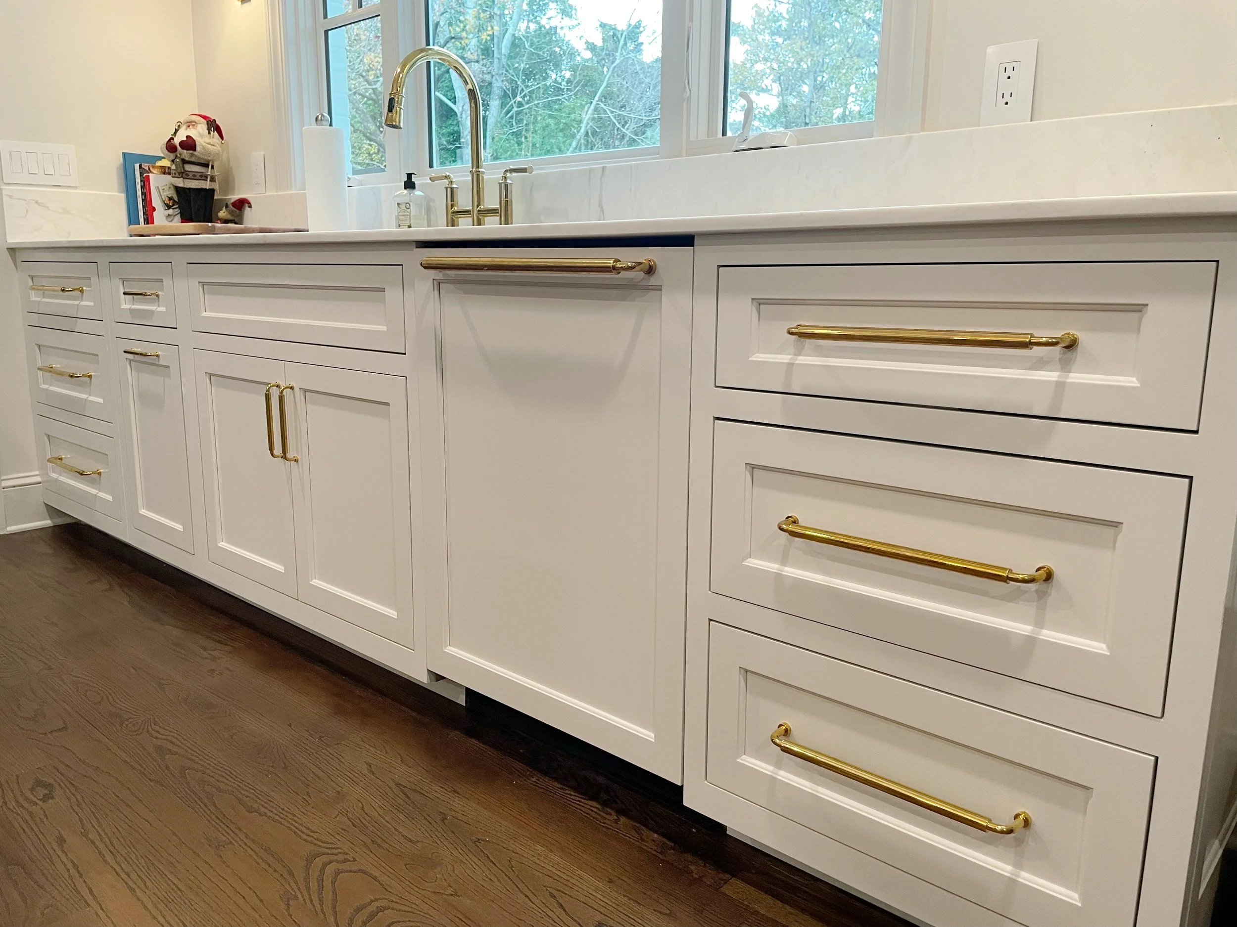 A white kitchen cabinet with gold handles and a gold towel bar below a window. There is a gold faucet over a white sink and a paper towel holder on the counter. Decor includes Christmas-themed items and books.