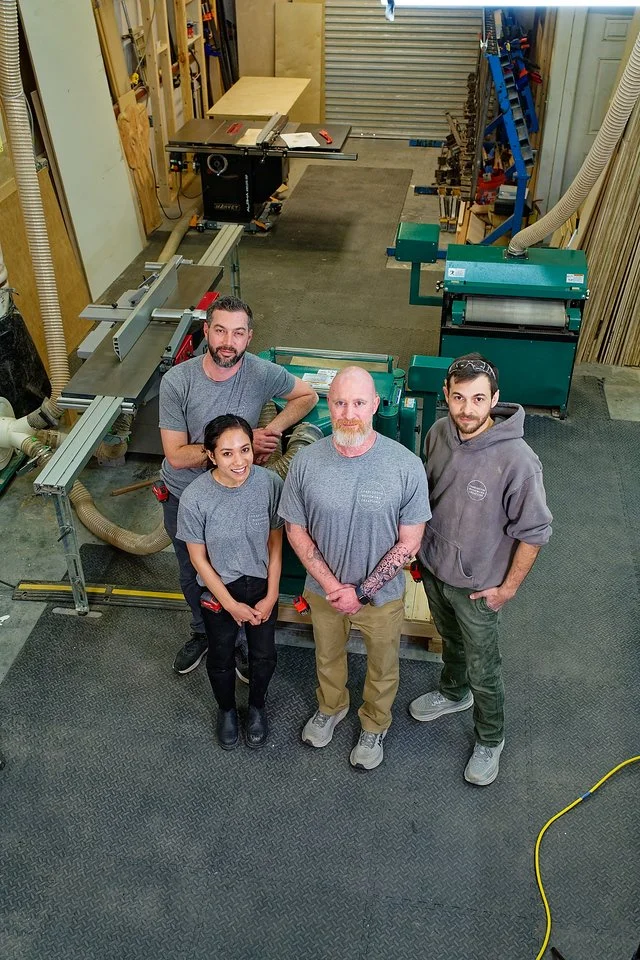 Five people standing in a woodworking shop surrounded by various tools and woodworking equipment, looking up at the camera.