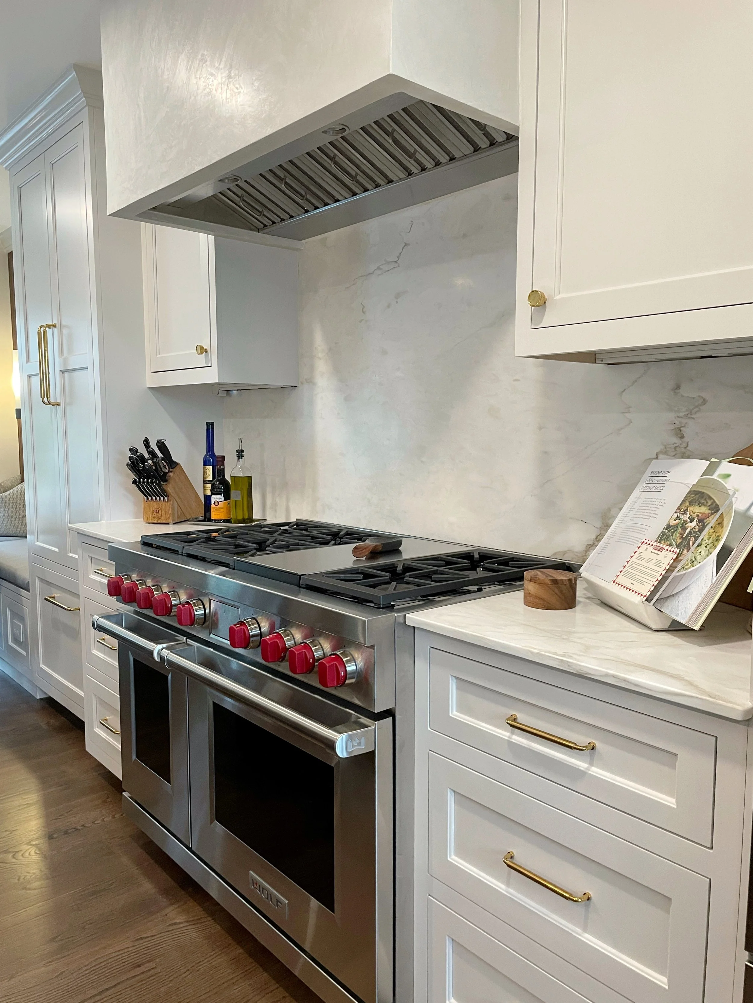 Modern kitchen with white cabinets, stainless steel oven, red knobs, a marble countertop, and cooking utensils, olive oil bottles, and a cookbook.