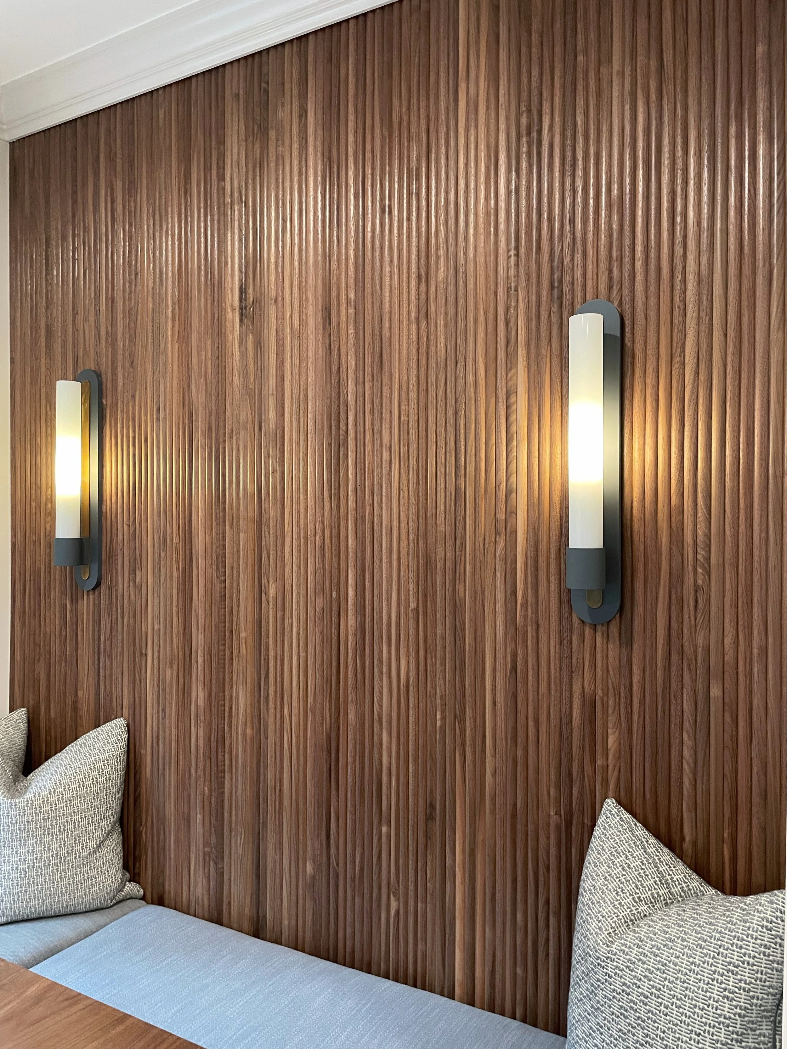 Wood-paneled wall with two modern wall sconces emitting warm light, and a part of a sofa with cushions visible at the bottom left.