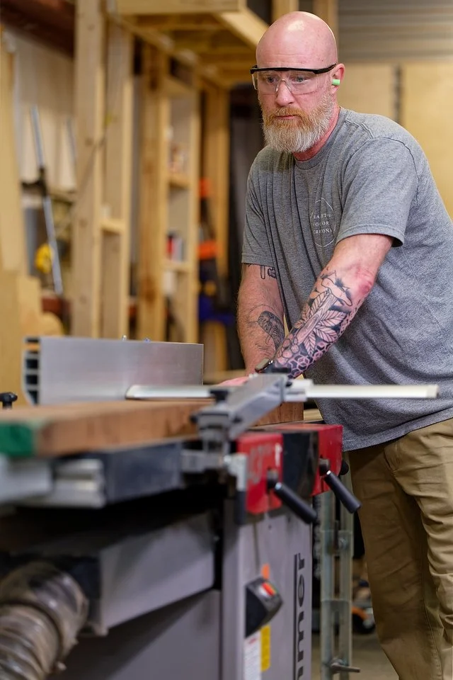A man with a beard, tattoos, safety glasses, and ear protection working in a woodworking shop, standing next to a table saw.