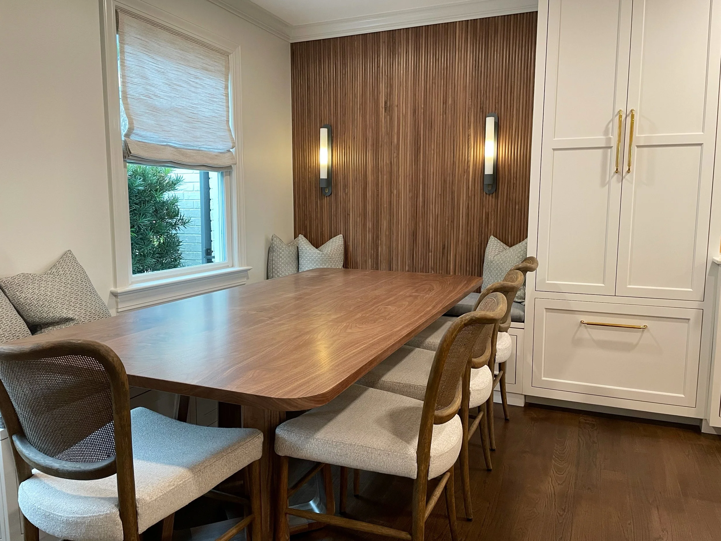 Dining room with a wooden table, upholstered chairs, a window with a shade, a wooden accent wall with wall sconces, and white cabinetry with gold handles.