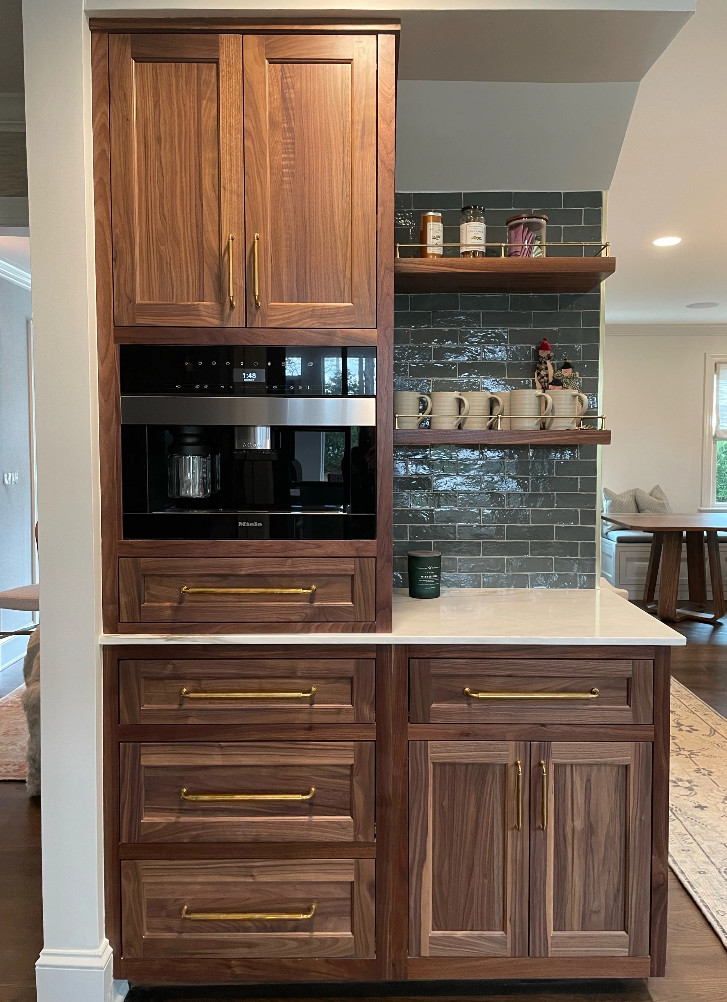 A kitchen built-in wooden cabinetry with brass handles, a built-in coffee machine, and a countertop. Open shelves above hold jars, mugs, and small decorative items. A living area is visible in the background.