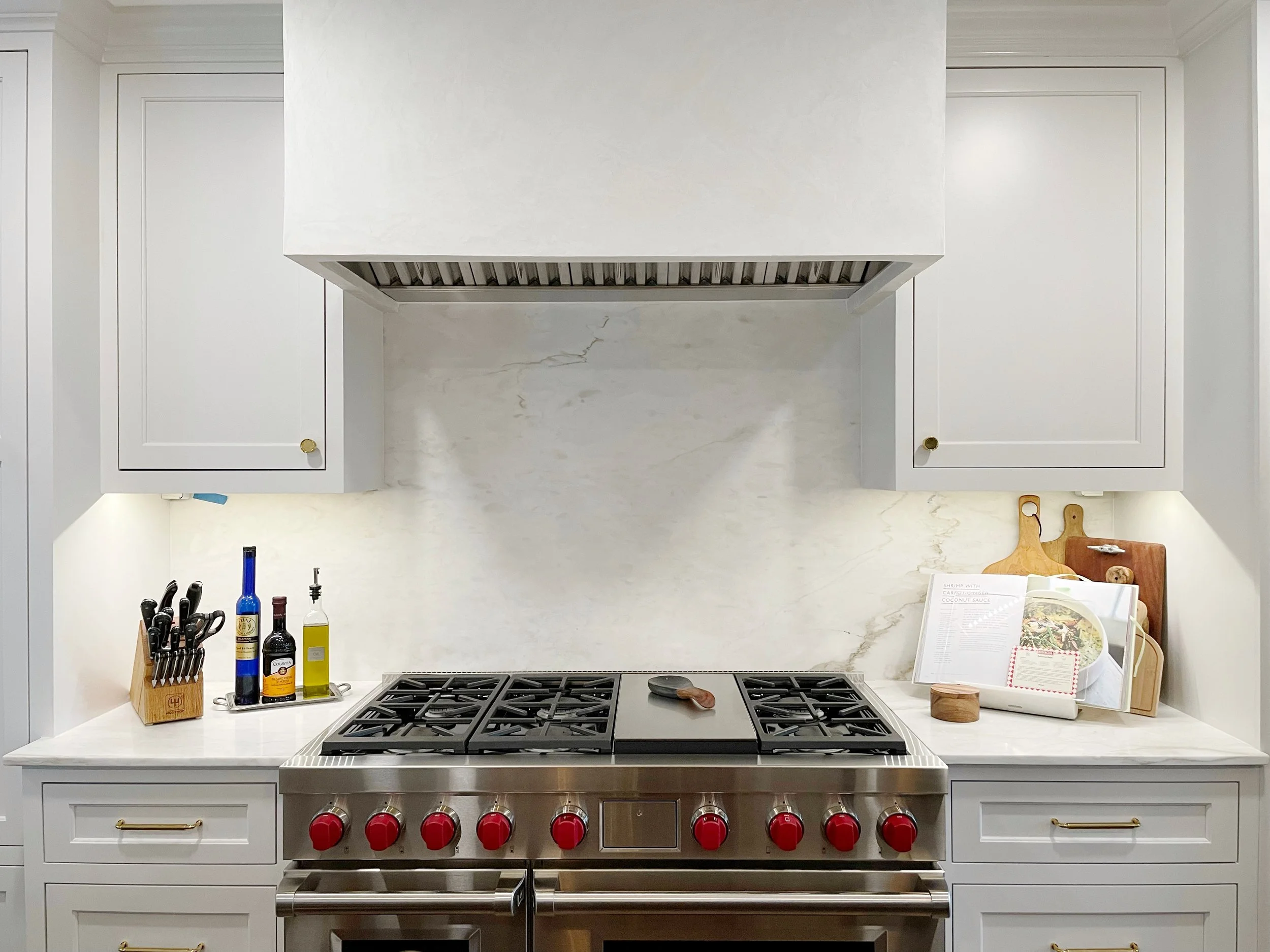 A modern kitchen with white cabinetry, a stainless steel stove with red knobs, and a white marble backsplash. There are kitchen utensils and bottles on the counter, along with cutting boards and a cookbook.