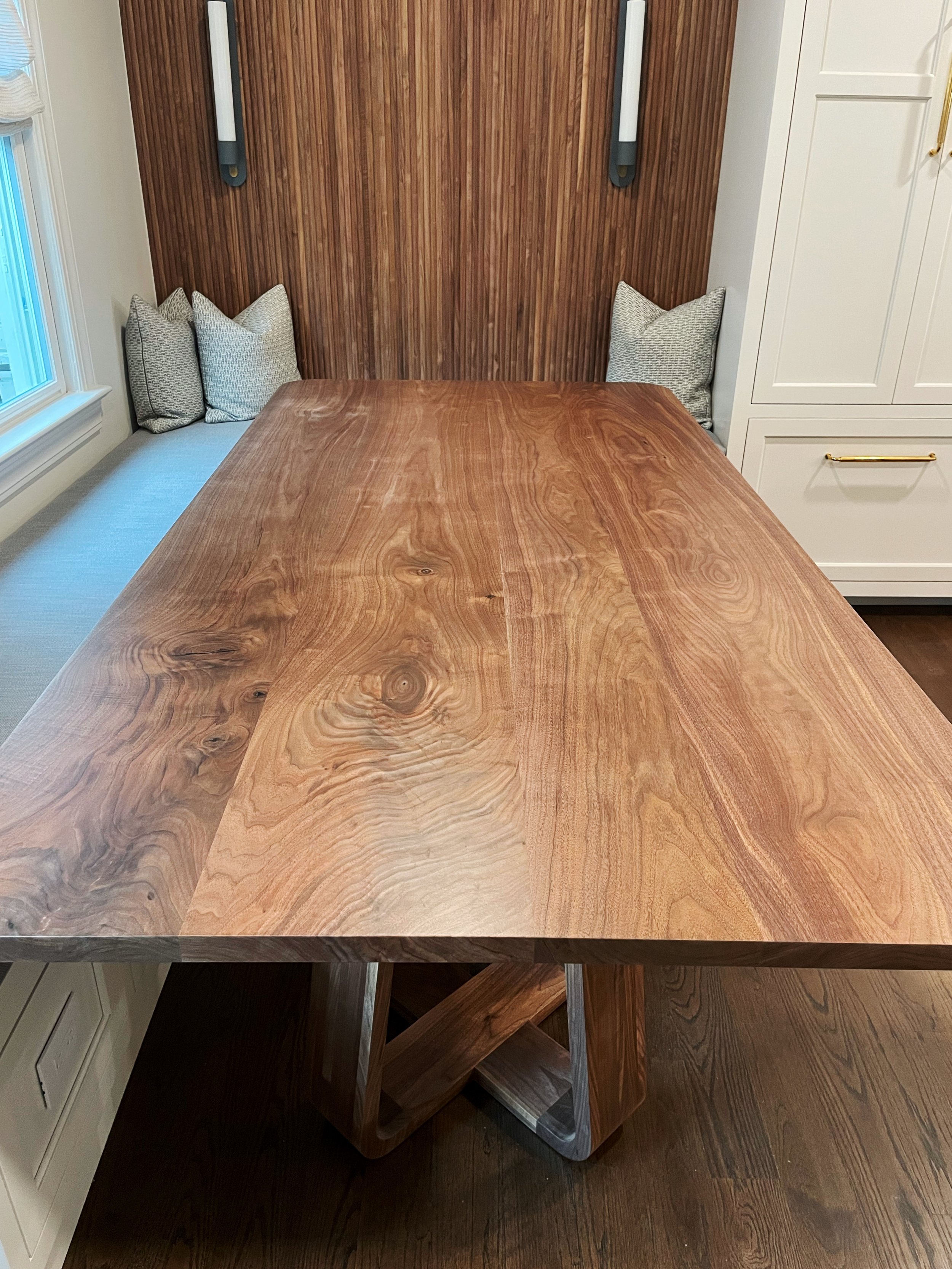 A wooden dining table with a natural finish and a unique base, set in a light-filled room with built-in white cabinets and patterned cushions on a bench along the wall.