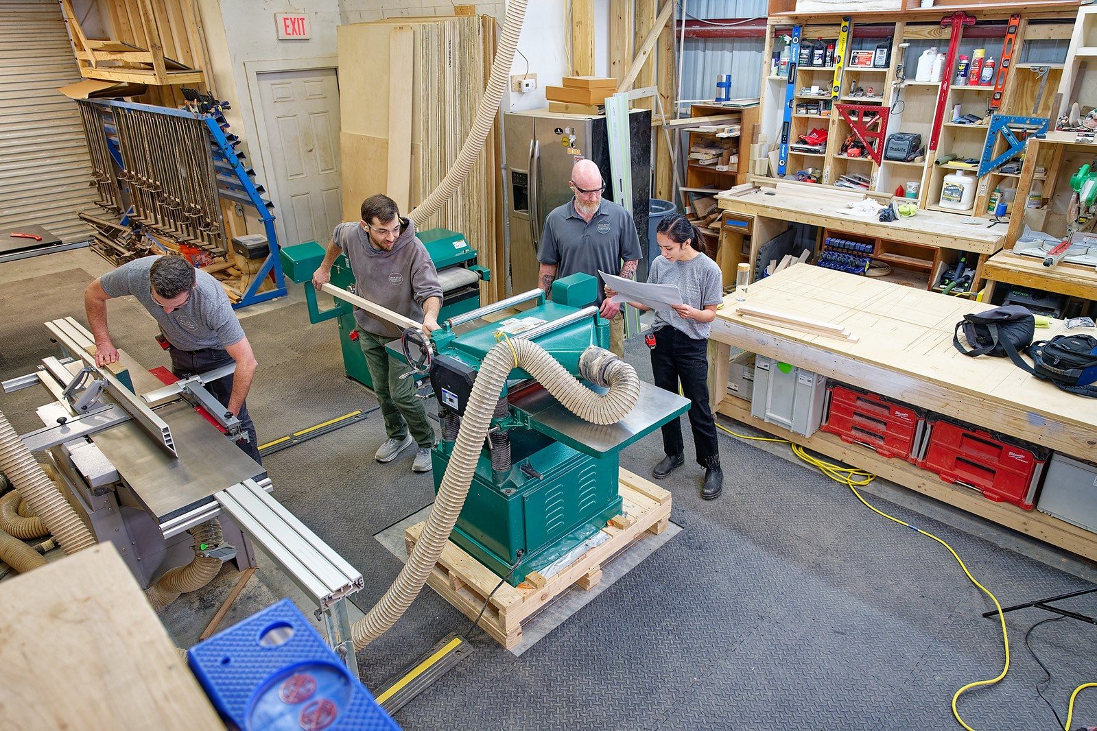 Four people working in a woodworking shop. One person is using a table saw, two are operating a large green machine, and one is reading a blueprint. The shop has various woodworking tools, lumber, and storage shelves along the walls.