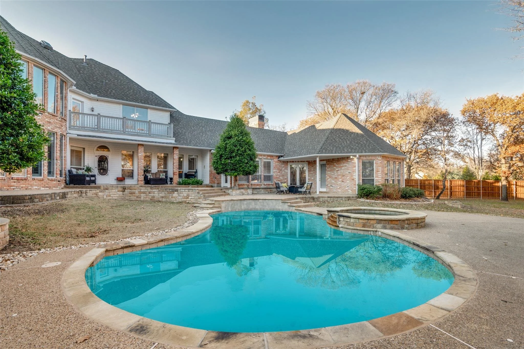 Large house with brick exterior featuring a backyard swimming pool, hot tub, patio area, and outdoor seating. Surrounded by trees and a wooden fence.