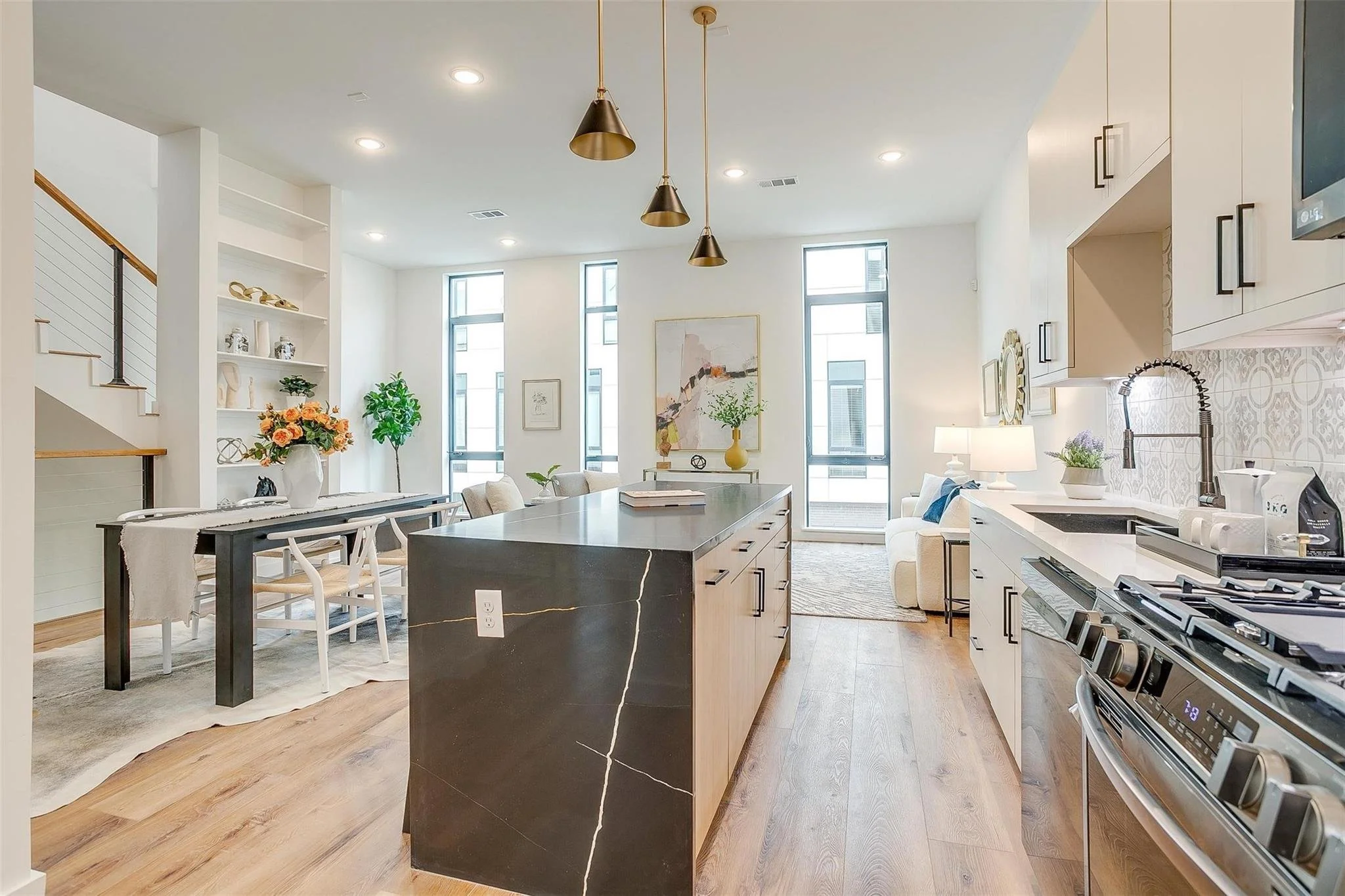 Modern open-concept kitchen and living area with a large kitchen island, pendant lighting, dining table with chairs, and decorative vases. Light wood flooring, white walls, high windows, and contemporary decor.