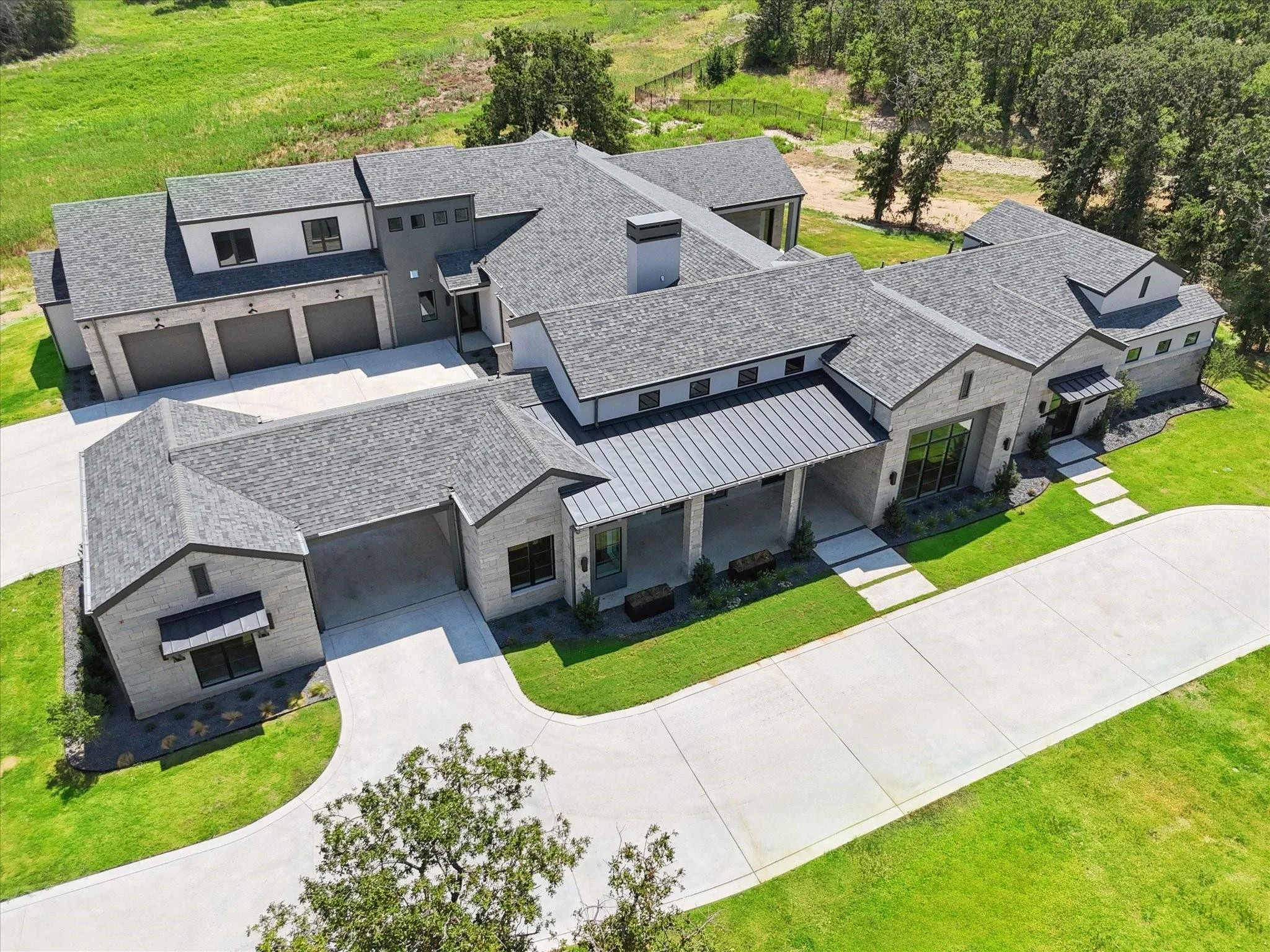 Aerial view of a large, modern suburban house with grey roofing and stone exterior, surrounded by a green lawn and a driveway.