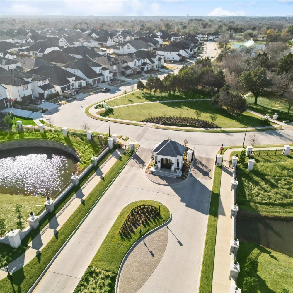 Aerial view of a residential neighborhood with a gated entrance, featuring landscaped roads and a small pond. Upscale houses are visible in the background.