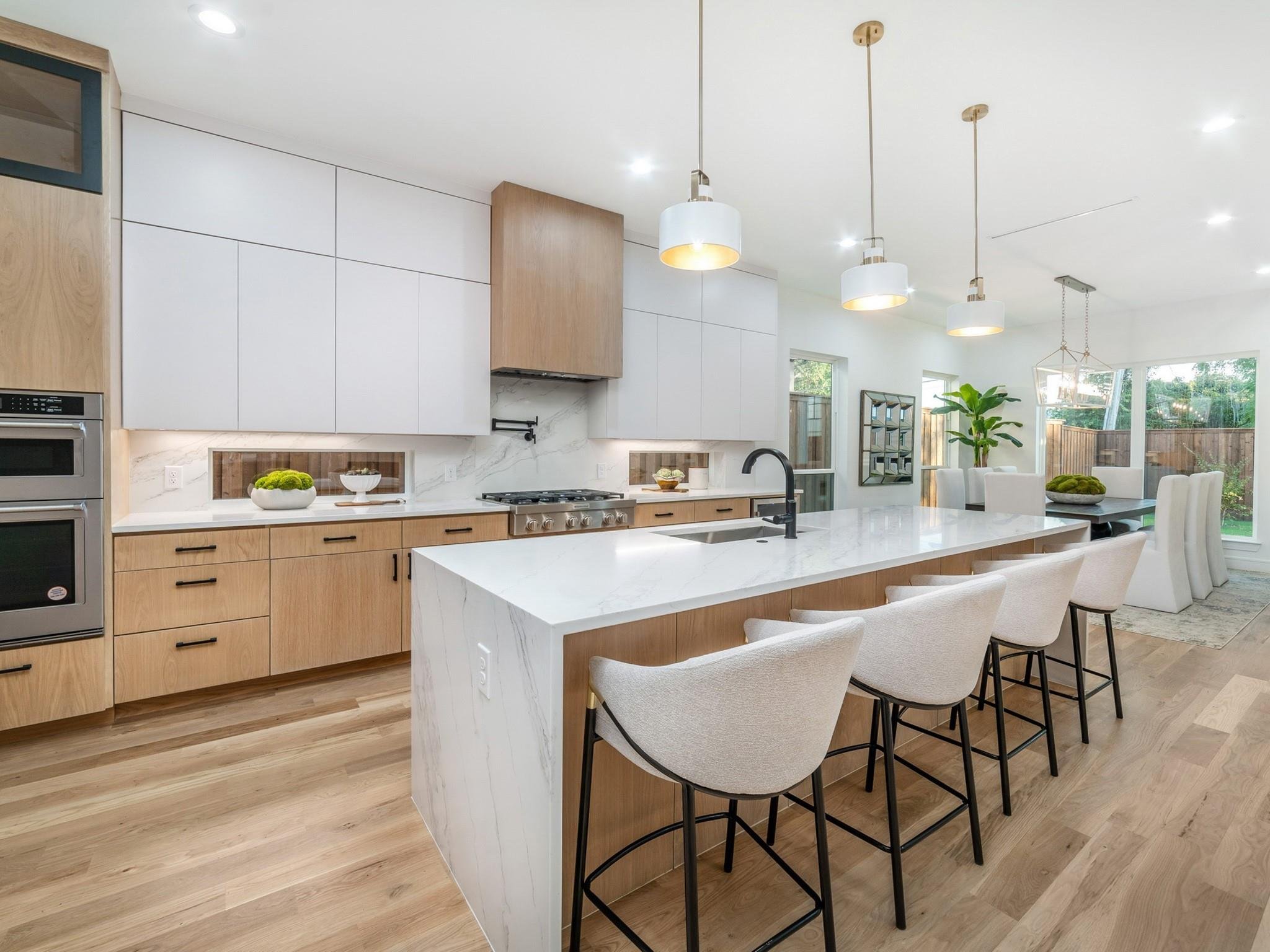 Modern kitchen interior with white cabinets, wooden drawers, marble countertops, and island with bar stools. Pendant lights hang above the island. Kitchen appliances include a built-in oven. The dining area is visible with a large window and plants.