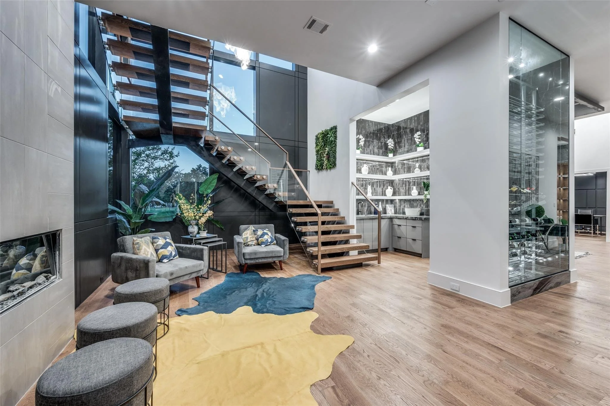 Modern interior with a wooden staircase, glass railing, and hardwood floors. Features a seating area with armchairs and a decorative rug, adjacent to a display shelf and a glass wine storage area.