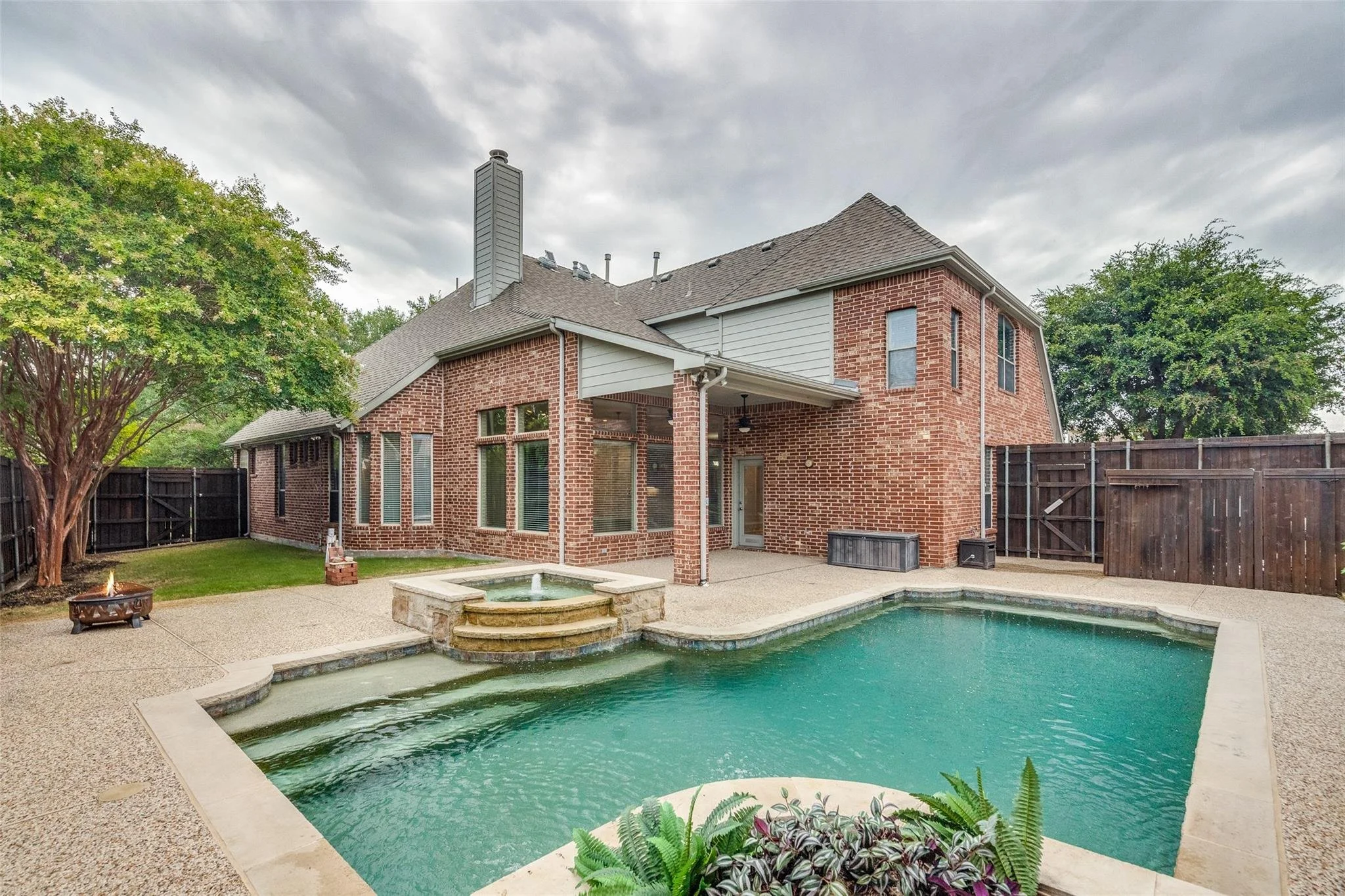 Brick house with backyard pool, hot tub, and patio. Tall windows and trees visible. Wooden fence surrounds the yard.