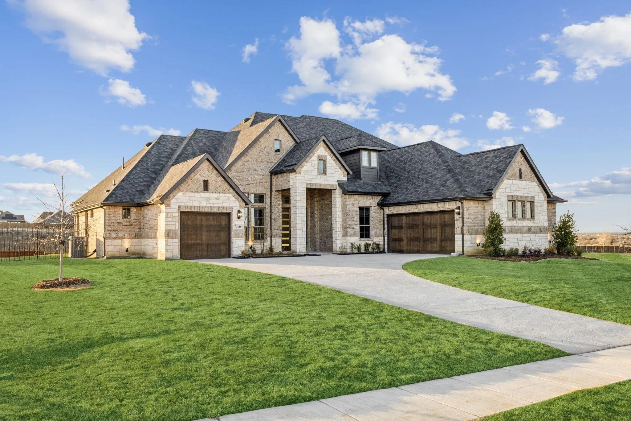 Large brick house with a dark shingled roof, three-car garage, and well-maintained lawn under a blue sky with clouds.