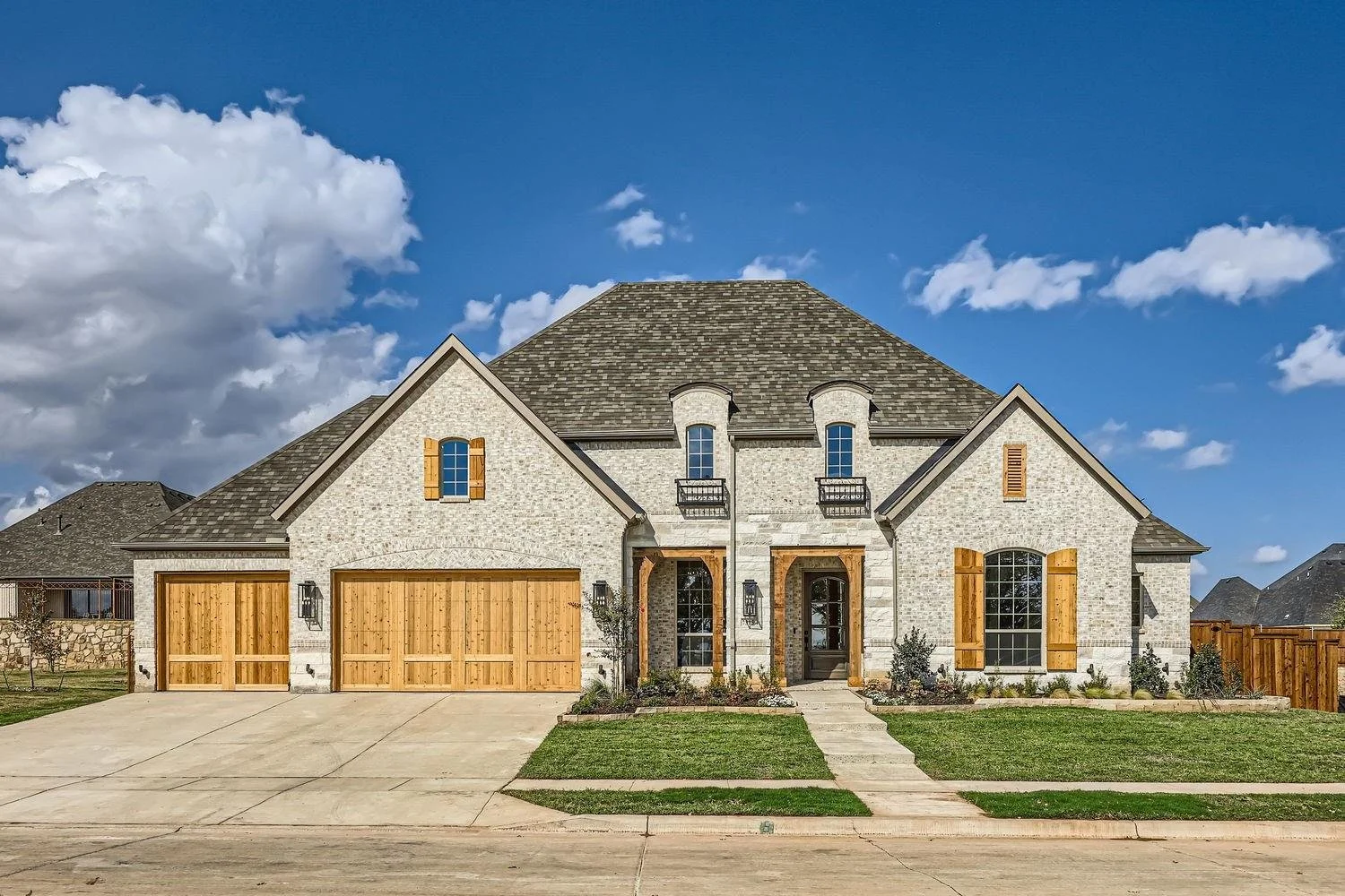 Two-story brick house with a three-car garage, wooden shutters, and a manicured lawn under a blue sky with clouds.