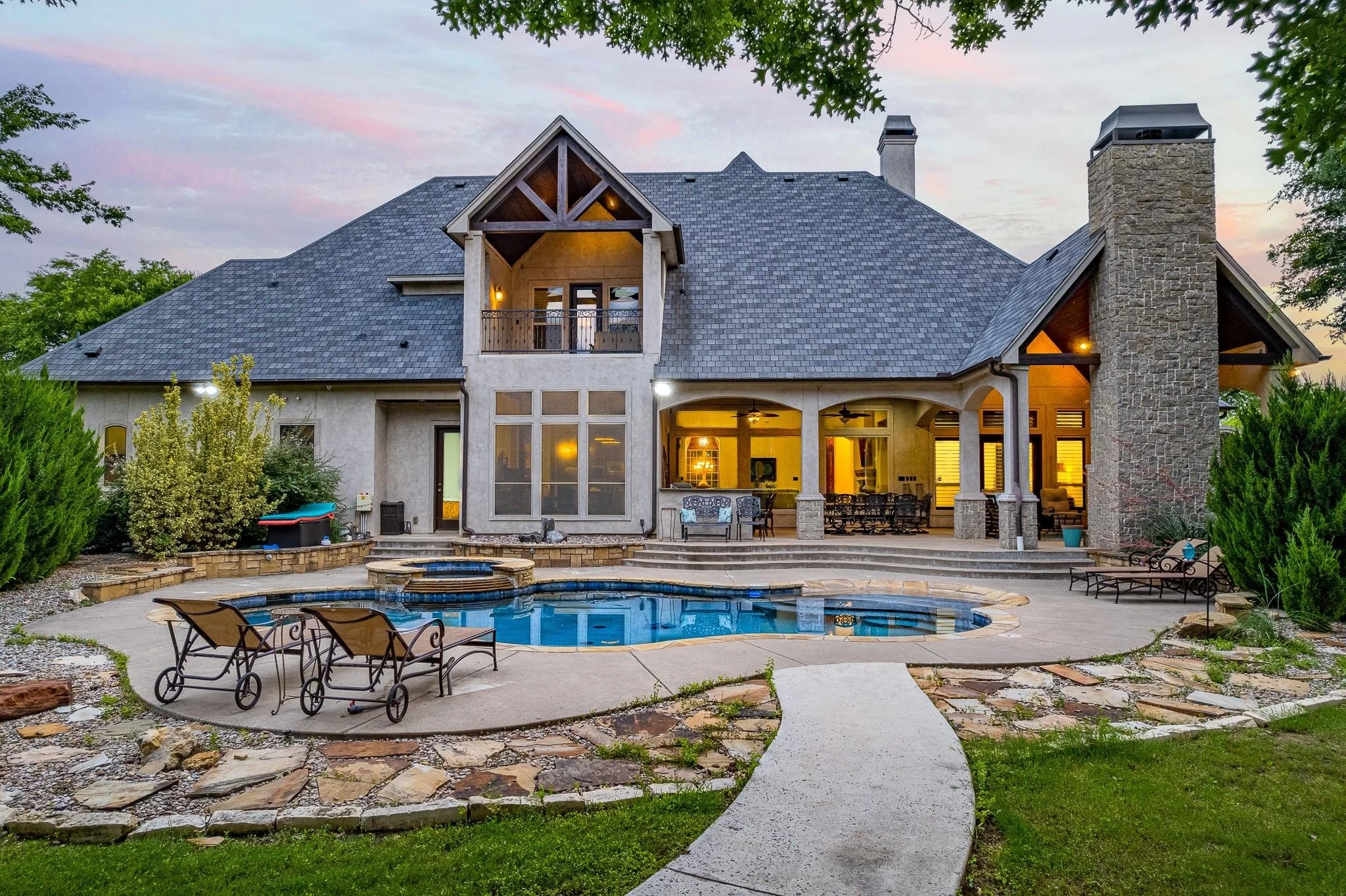 Luxurious house with a swimming pool, patio area, stone chimney, and landscaped garden at sunset.