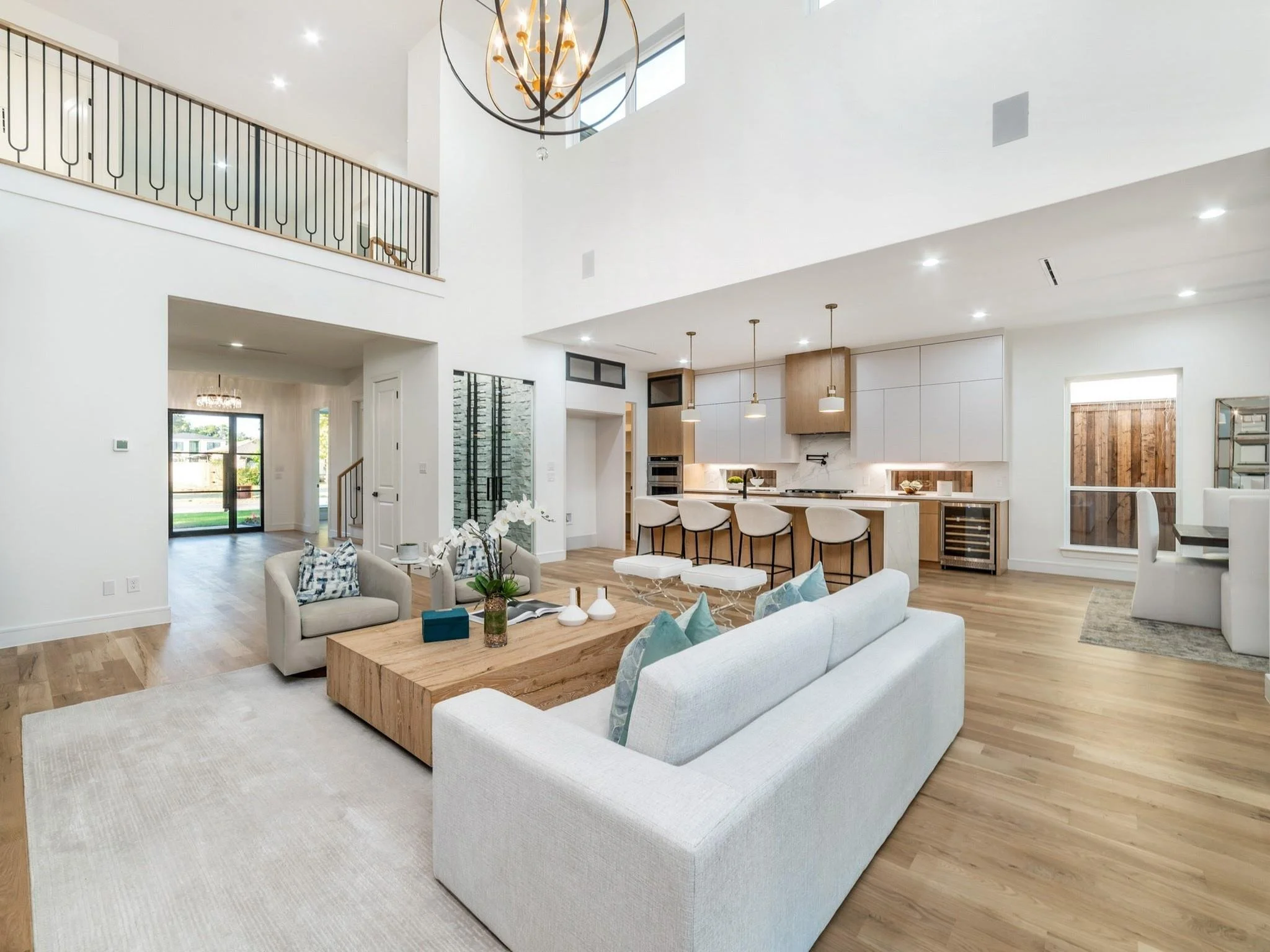 Modern open-concept living space with high ceilings, featuring a living room with white sofas and a wooden coffee table, a kitchen with a large island and bar stools, and a dining area. Light wood flooring and a contemporary chandelier are present.
