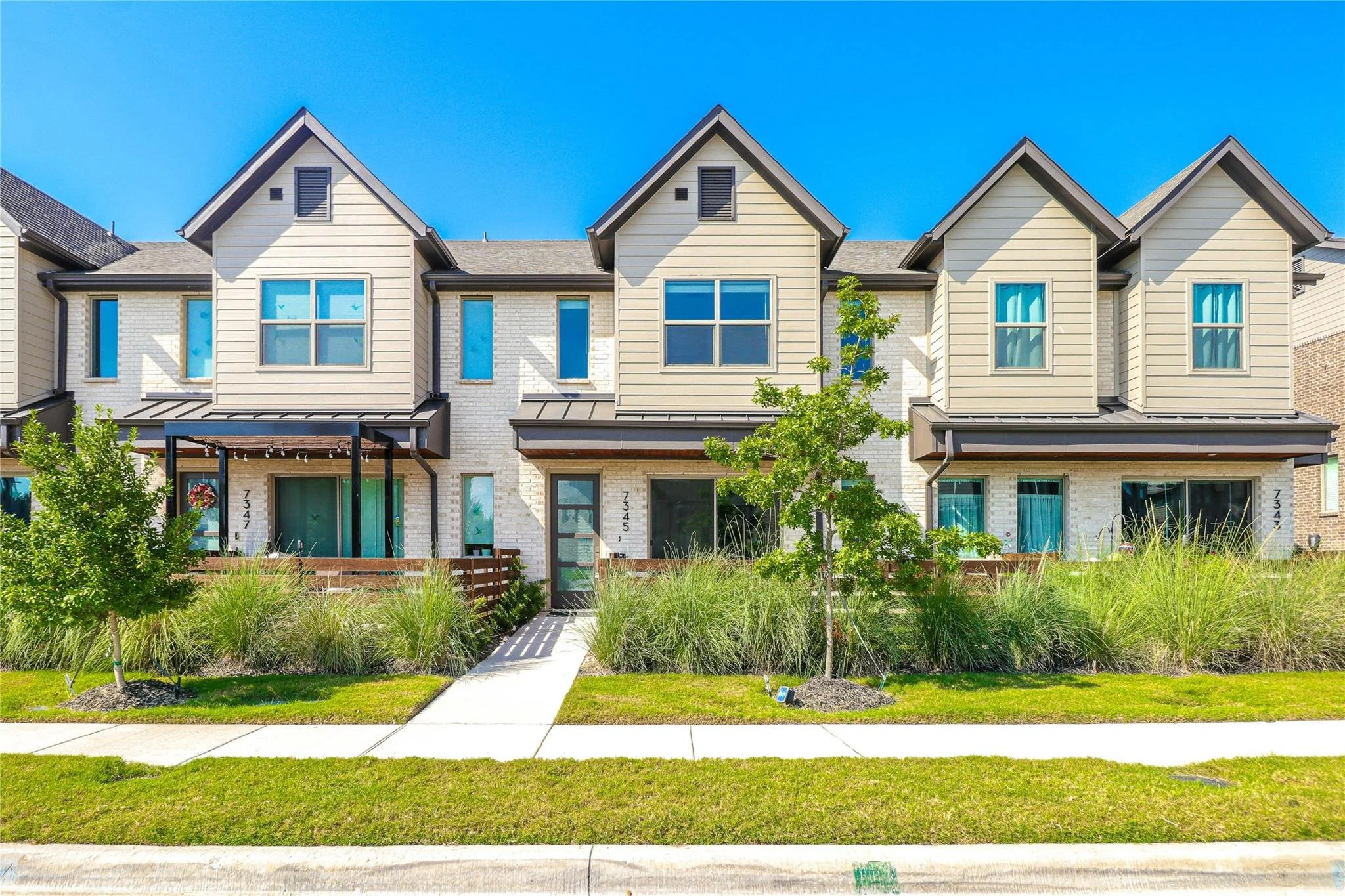 Modern townhouse exterior with beige siding, brown trim, landscaped front yard, and blue sky background