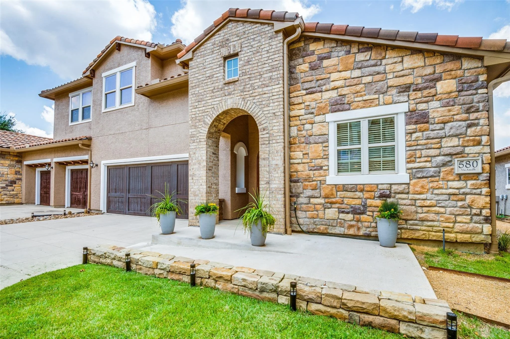 Modern two-story house with stone and stucco exterior, featuring a double garage, large arched entrance, and potted plants lining the pathway.